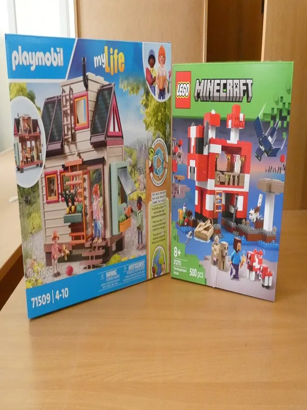Dwa pudełka z klockami na drewnianym stole. Po lewej stronie pudełko Playmobil z serii myLife (model 71509, 160 elementów, wiek 4-10 lat), przedstawiające dom z postaciami ludzkimi, drzewami i detalami ogrodu. Po prawej stronie pudełko LEGO Minecraft (model 21179, 500 elementów, wiek 8+), ukazujące czerwono-białą konstrukcję z wodą, postaciami z gry i smokiem. Na obu pudełkach widoczne są logotypy, liczby elementów oraz informacje wiekowe. Tło stanowi drewniana szafka i okno.