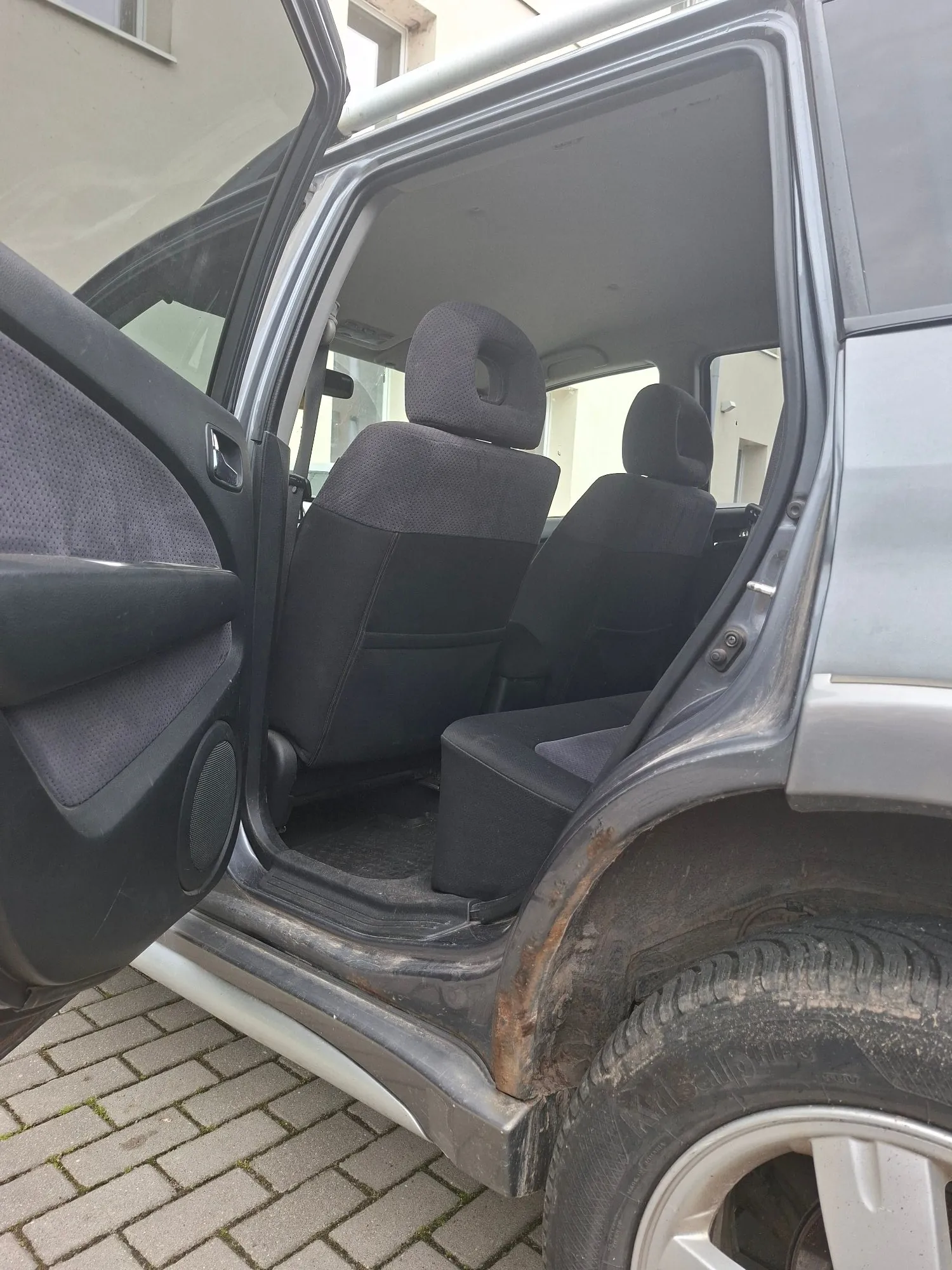 Zdjęcie wnętrza samochodu z otwartymi drzwiami, ukazujące czarne materiałowe fotele. Widoczna jest korozja na progu nadwozia w okolicach zawiasów drzwi. W prawym górnym rogu widoczny jest fragment koła z oponą i felgą, a w tle kostka brukowa.
