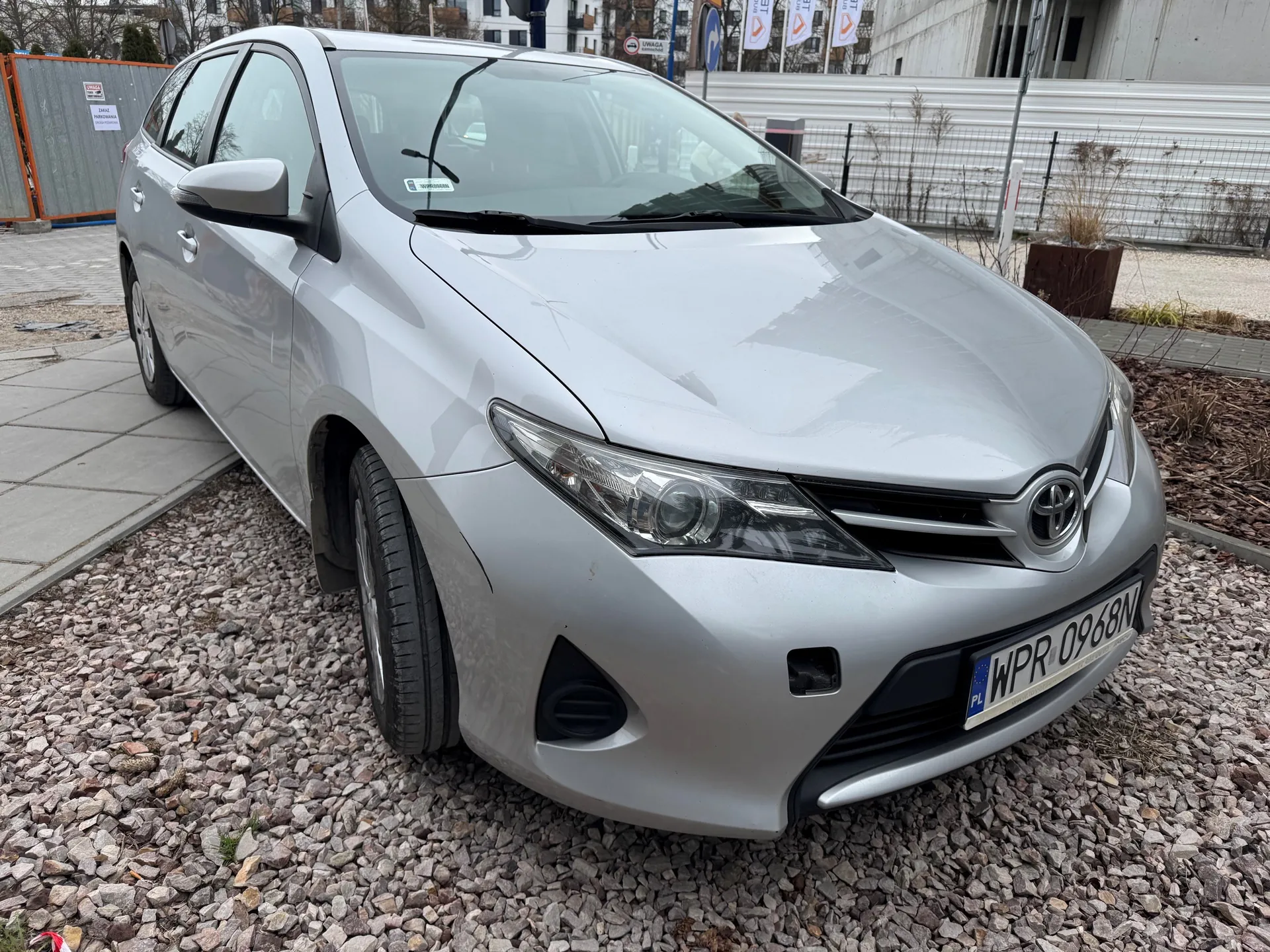 Srebrny samochód osobowy marki Toyota zaparkowany na podłożu z żwiru. Widoczna tablica rejestracyjna PL WPR 0968N. W tle budynki, metalowe ogrodzenie, barierki budowlane oraz znaki drogowe.