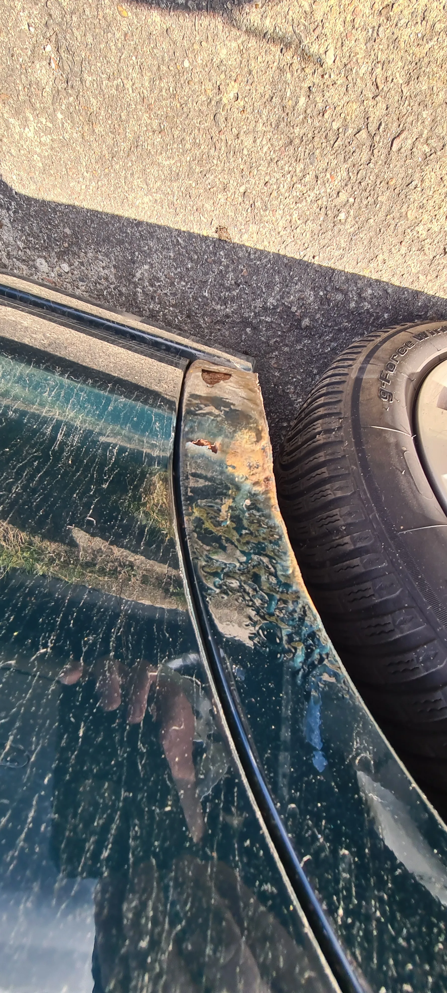 Zbliżenie na uszkodzony fragment nadwozia samochodu w kolorze ciemnoniebieskim. Widoczne są zarysowane ślady korozji i obłupionej farby na zderzaku. Koło z oponą marki g-Force, na której widnieje napis. Szkło boczne z widocznymi pionowymi zadrapaniami i odbiciem dłoni trzymającej aparat. Tło stanowi asfaltowa powierzchnia i fragment betonowego chodnika.