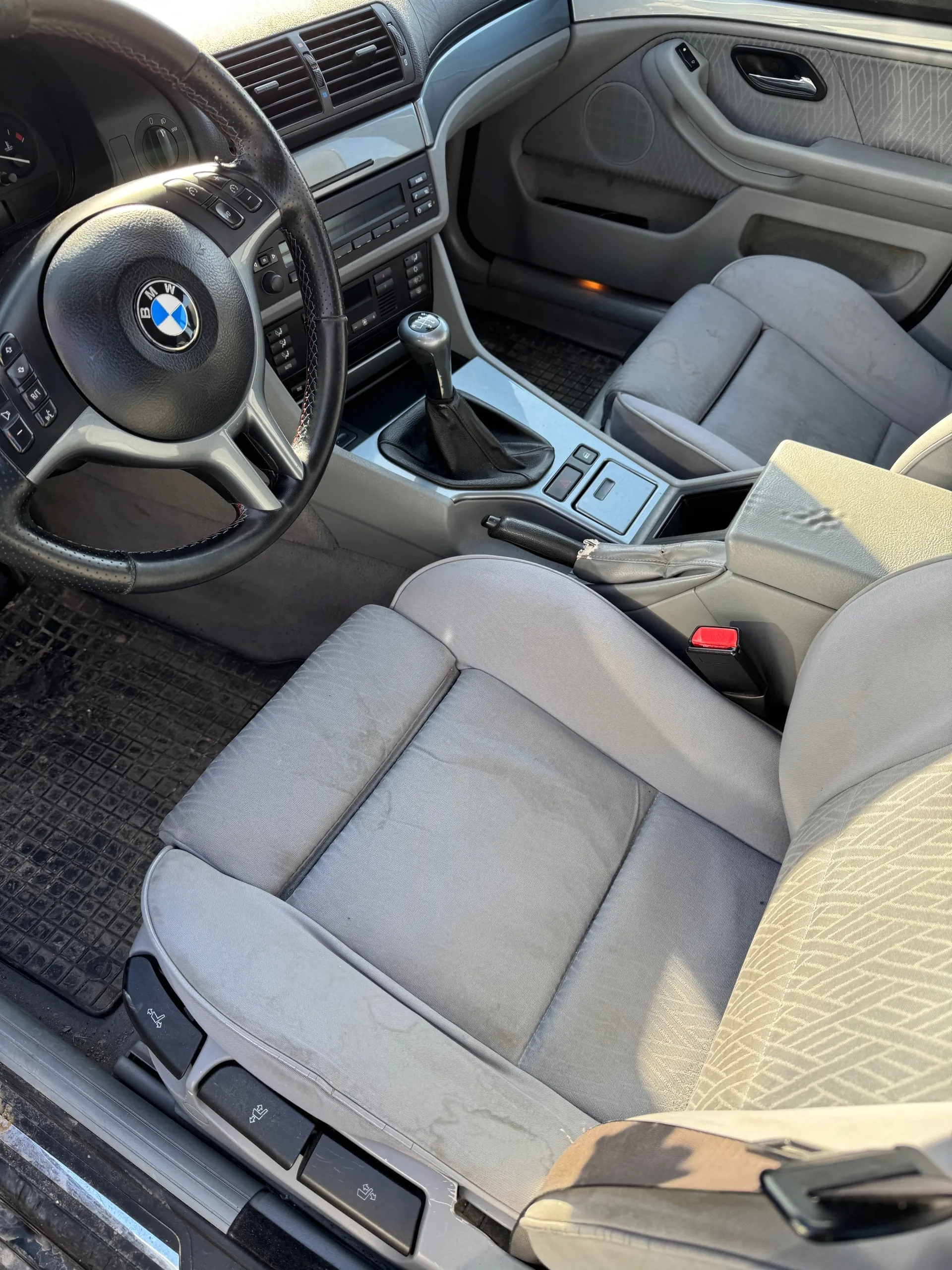 Wnętrze samochodu marki BMW z widocznym kierownicą z logo, szarymi siedzeniami tapicerowanymi tkaniną, centralną konsolą z dźwignią zmiany biegów, czarnymi dywanikami podłogowymi oraz panelami drzwiowymi z głośnikami. Na kierownicy znajdują się przyciski sterujące, a na desce rozdzielczej widoczne są wentylacyjne otwory i system audio. Siedzenia mają lekkie ślady zużycia, a wewnątrz pojazdu panuje oświetlenie słoneczne.