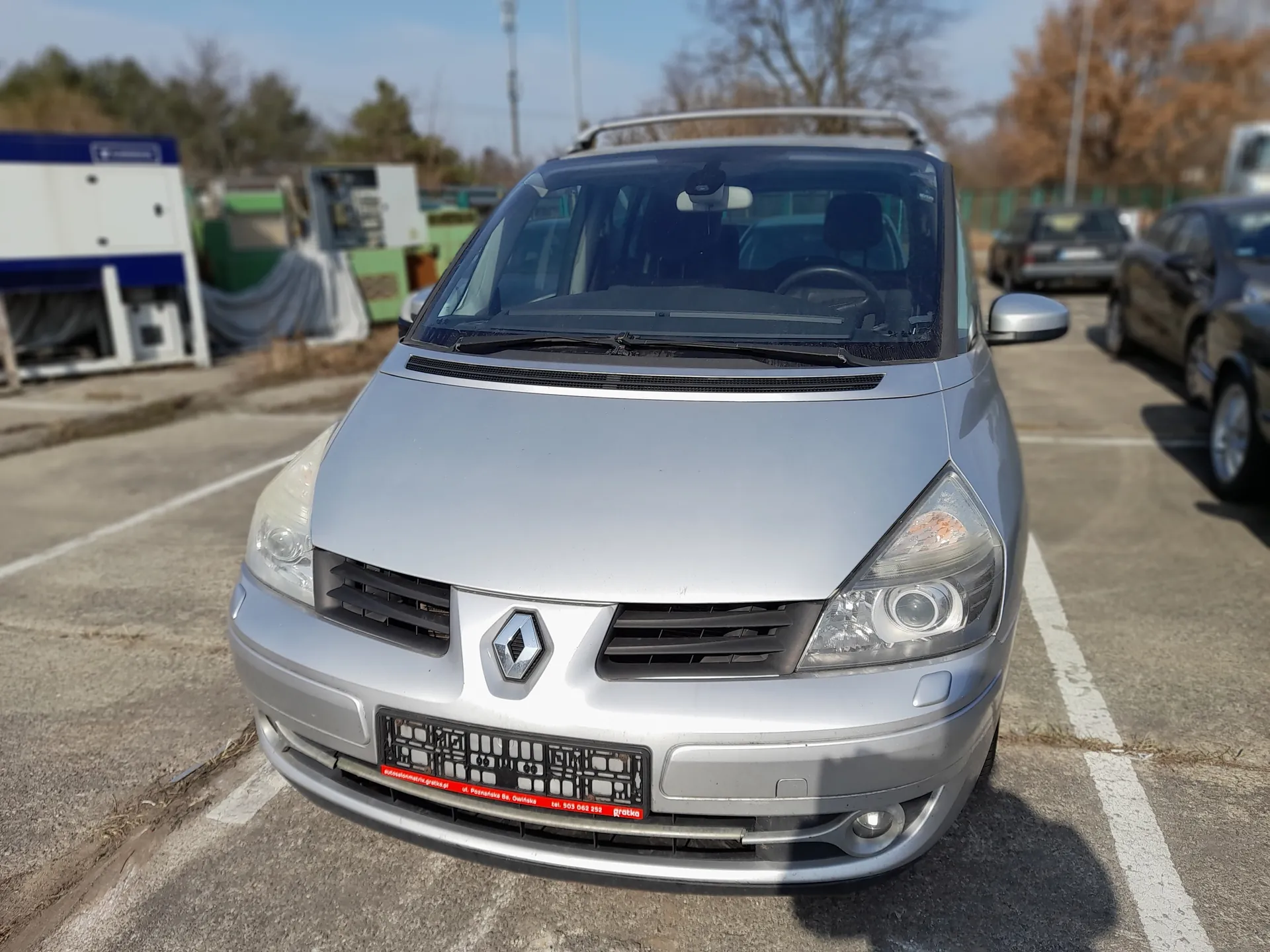 Srebrny samochód marki Renault zaparkowany na placu parkingowym, widok z przodu. Na masce widoczne logo Renault, tablica rejestracyjna z czerwonym napisem. W tle inne pojazdy, budynki, drzewa oraz jasne niebo.