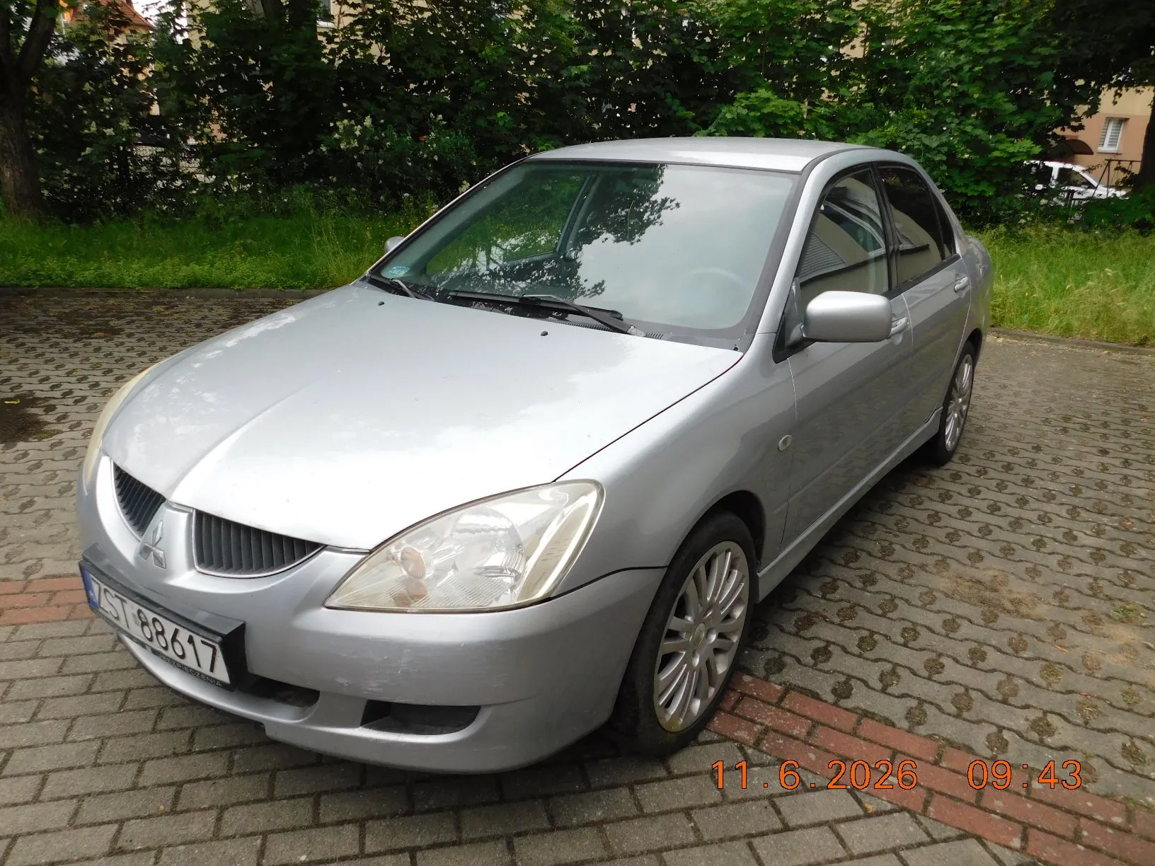 Srebrny samochód osobowy marki Mitsubishi zaparkowany na kostce brukowej. W tle widoczne są zielone krzewy i drzewa. Na przednim zderzaku znajduje się tablica rejestracyjna o numerze ZST 88617. W prawym dolnym rogu zdjęcia widnieje pomarańczowy znacznik daty i godziny: 11. 6. 2026 09:43.