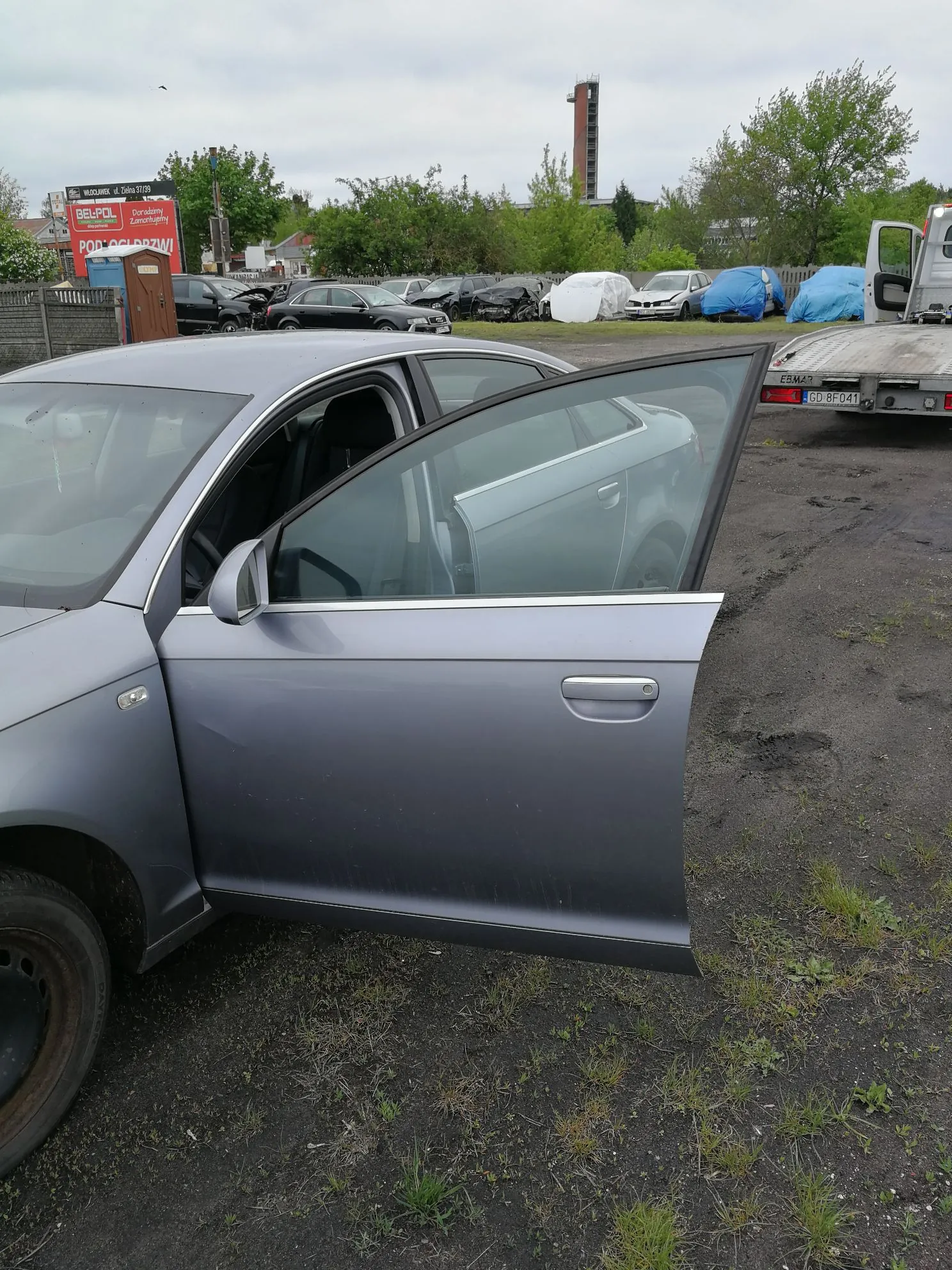 Zdjęcie przedstawia szary samochód osobowy z otwartymi przednimi drzwiami od strony kierowcy. Pojazd znajduje się na placu z ciemnej ziemi i żwiru. W tle widoczne są inne samochody, w tym niektóre przykryte niebieskimi i białymi plandekami, oraz tablica reklamowa z napisem 'BELPOL' i 'POD OCHRONĄ!'. W oddali widać zielone drzewa oraz wysoką, wąską konstrukcję przypominającą komin lub wieżę na tle zachmurzonego nieba.