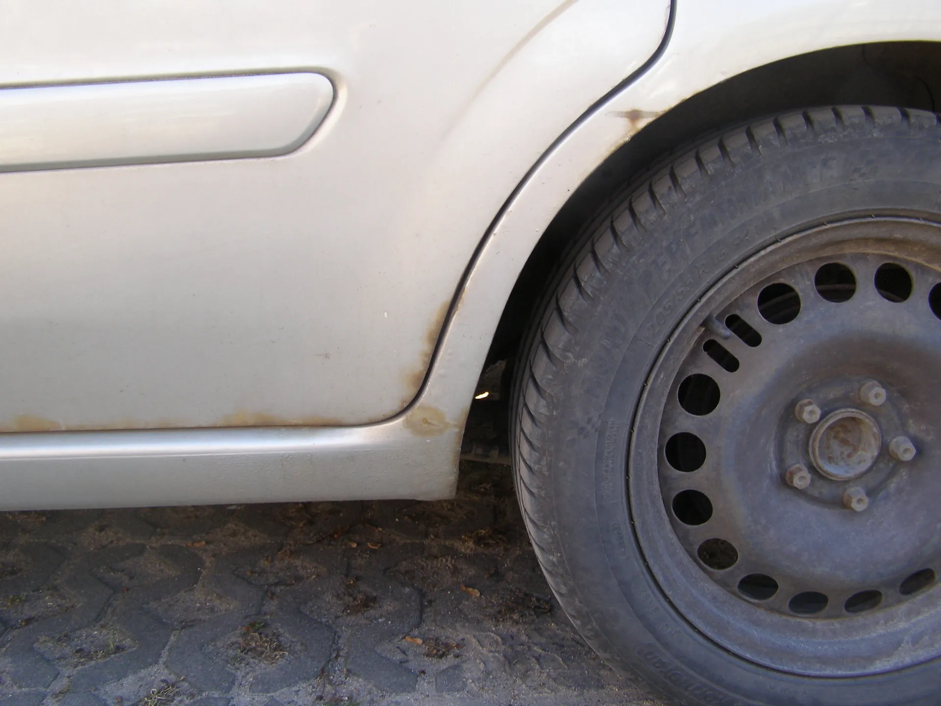Zbliżenie na tylną część białego samochodu, obejmujące koło i fragment karoserii. Widoczne są oznaki rdzy na karoserii w okolicy nadkola oraz na obręczy koła. Opona ma czarną powierzchnię z widocznymi rysami i napisami, a felga posiada otwory oraz zardzewiałe śruby. Pod samochodem znajduje się brukowana powierzchnia z drobnymi plamami ziemi i trawy.