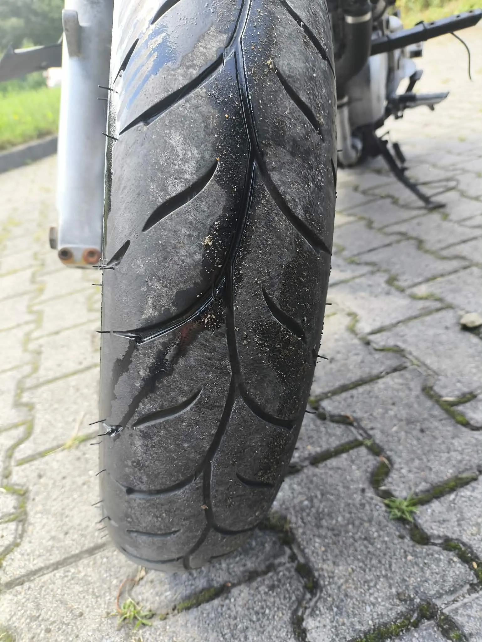 Zbliżenie na przednią oponę motocyklową z widocznym bieżnikiem, ustawioną pionowo na szarym, kostkowym podłożu. Opona jest lekko zabrudzona i wilgotna.