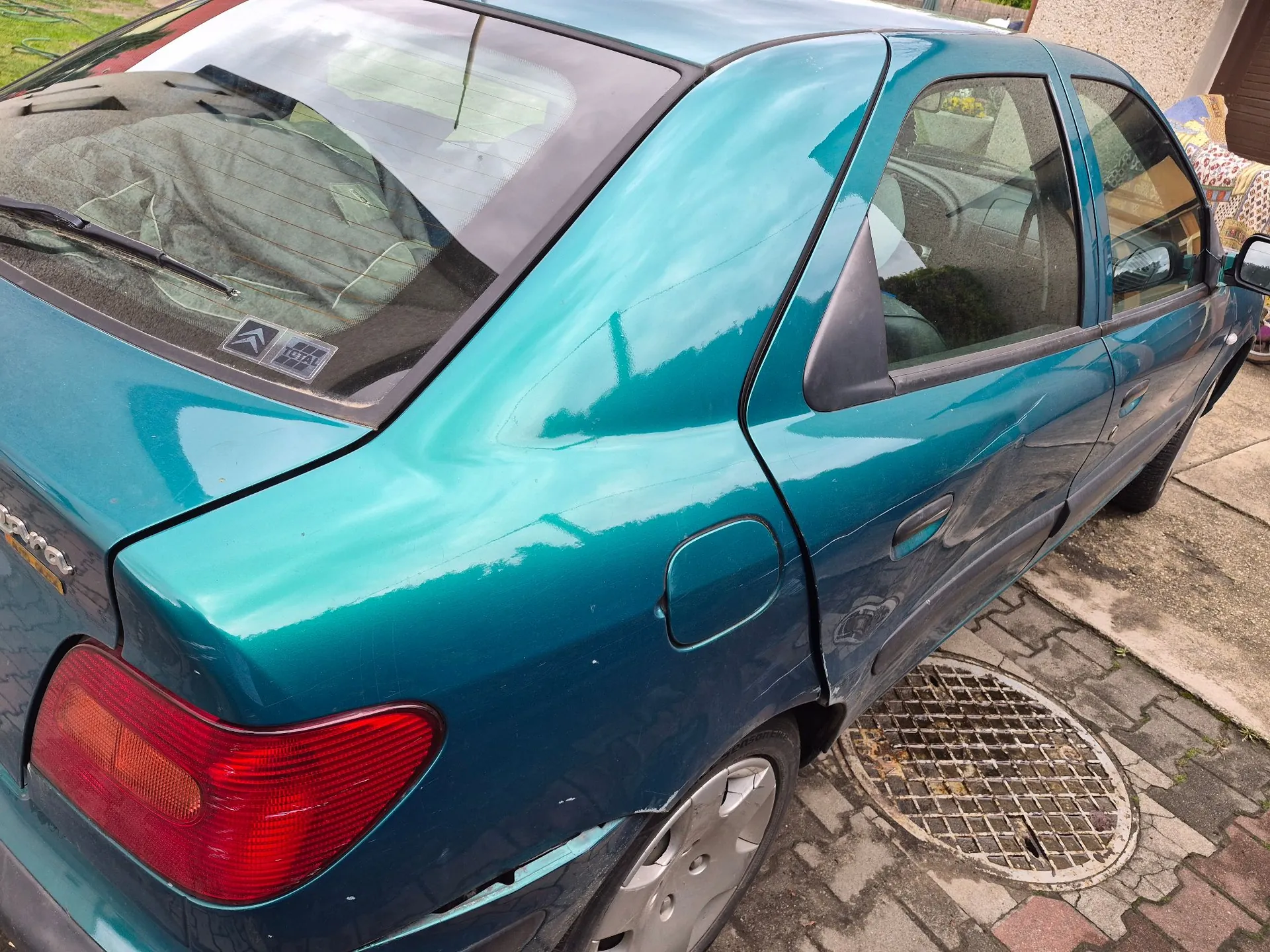 Zdjęcie tylnej części i boku samochodu w kolorze ciemnoturkusowym. Widoczna jest tylna szyba z wycieraczką i logo Citroën, klapka wlewu paliwa oraz tylne światło. Karoseria w dolnej części drzwi i nad nadkolem wykazuje ślady uszkodzeń i wgnieceń. Samochód zaparkowany jest na kostce brukowej obok metalowej kratki ściekowej.