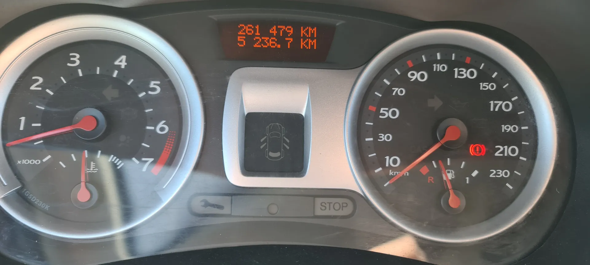 Tablica rozdzielcza samochodu z dwoma głównymi wskaźnikami. Po lewej stronie wskaźnik obrotomieru z czerwoną igłą wskazującą około 1,5 (skala do 7 x1000 obr./min), poniżej niego wskaźnik temperatury chłodniczka. Po prawej stronie prędkościomierz z czerwoną igłą na 0 km/h (skala do 230 km/h), poniżej wskaźnik paliwa. W centralnej części cyfrowy wyświetlacz z napisem "261 479 KM" i "5 236,7 KM", poniżej ikona samochodu z otwartymi drzwiami. Na dole przycisk z napisem "STOP" oraz czerwone ostrzeżenie w kształcie koła z wykrzyknikiem. Wskaźniki i tekst są prawidłowo zorientowane.
