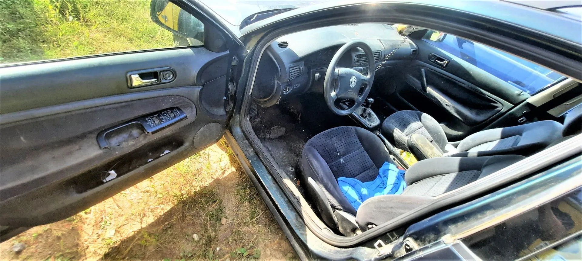 Wnętrze ciemnego samochodu z otwartymi drzwiami kierowcy. Widoczna jest kierownica z logo Volkswagena, deska rozdzielcza, dwa przednie fotele z ciemnej tkaniny, z czego jeden ma na siedzisku niebieską szmatkę. Drzwi kierowcy są szeroko otwarte, ukazując panel z klamką i przyciskami sterowania szybami. Samochód stoi na piaszczystym podłożu z trawą w tle.