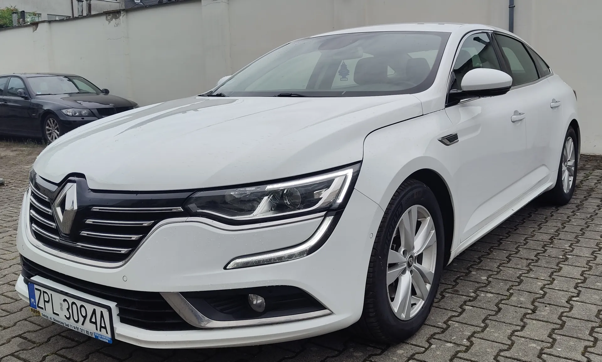 Biały samochód marki Renault Talisman zaparkowany na kostce brukowej. Widoczny jest przód i prawy bok pojazdu. Samochód ma polską tablicę rejestracyjną o numerze ZPL 3094A. W tle widoczny jest fragment ciemnego samochodu oraz jasna ściana budynku.