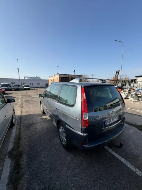 Srebrnoszary samochód marki Citroën w nadwoziu kombi zaparkowany na asfaltowym parkingu. Widoczny jest tył pojazdu z tablicą rejestracyjną GD 3H906. W tle znajdują się inne zaparkowane samochody, niskie budynki przemysłowe oraz latarnie uliczne pod bezchmurnym, niebieskim niebem.
