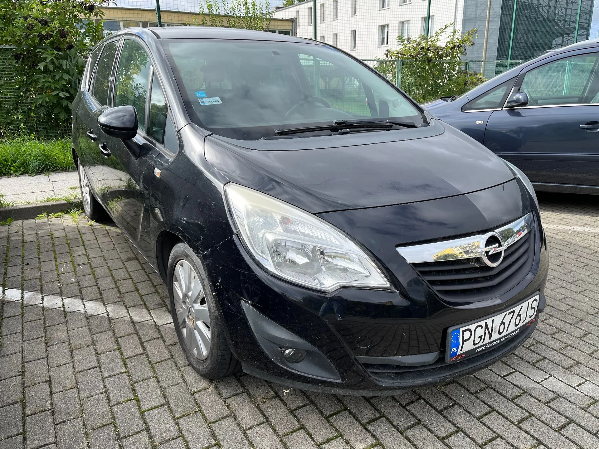 Czarny samochód osobowy marki Opel Zafira zaparkowany na kostce brukowej. Pojazd jest widoczny z przodu i z boku. Na przednim zderzaku znajduje się polska tablica rejestracyjna o numerze PGN 676JS.