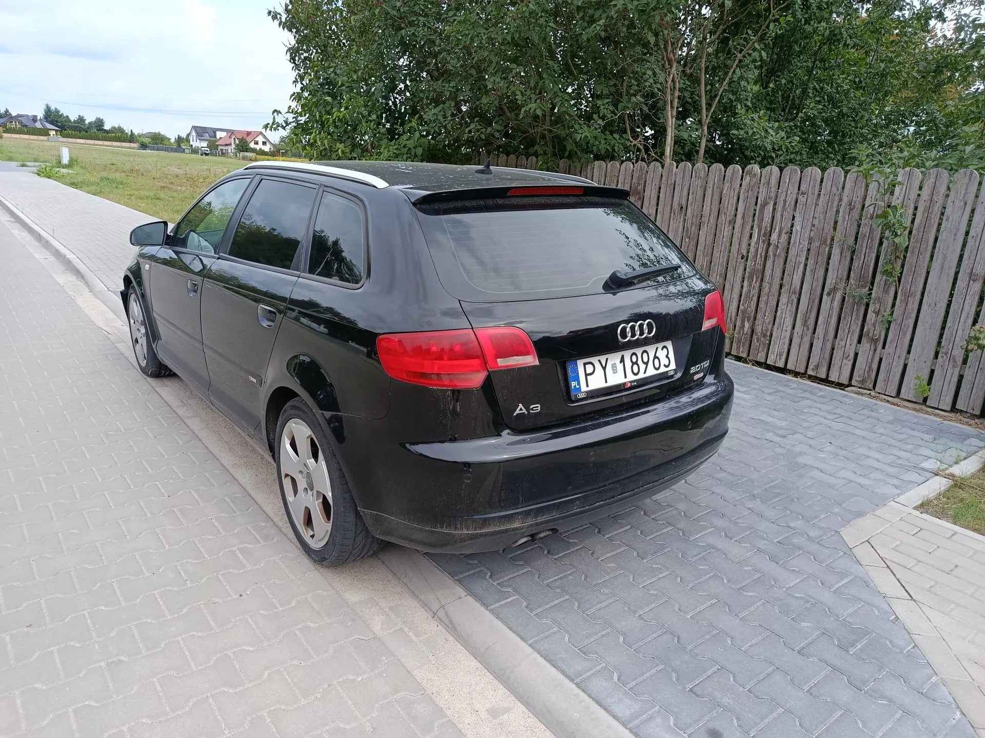 Czarny samochód marki Audi A3 Sportback zaparkowany na kostce brukowej obok krawężnika. Pojazd jest widoczny od tyłu i z boku. Na klapie bagażnika znajduje się logo Audi, oznaczenie modelu A3 oraz polska tablica rejestracyjna o numerze PY 18963. W tle widoczny jest drewniany płot, zielone drzewa oraz fragment trawnika i domów w oddali pod jasnym niebem.