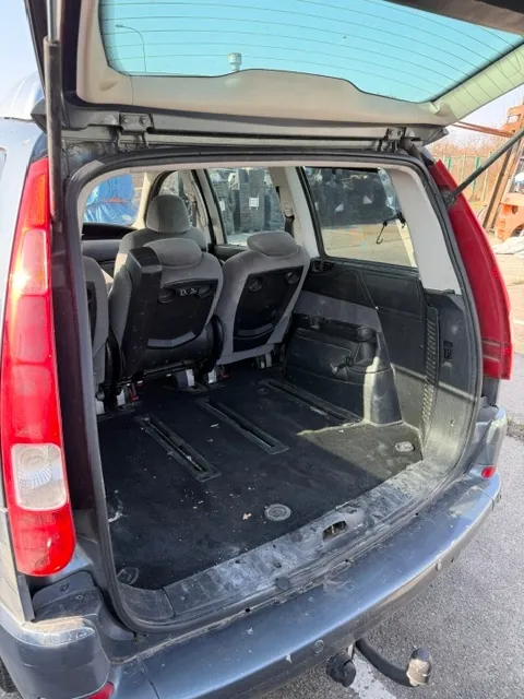 Widok z tyłu do otwartego bagażnika ciemnoszarego samochodu typu kombi lub minivan. Wewnątrz widoczne są złożone oparcia tylnych siedzeń oraz czarna wykładzina bagażnika. Po prawej i lewej stronie znajdują się pionowe czerwone lampy tylne. Samochód posiada hak holowniczy zamontowany pod zderzakiem.