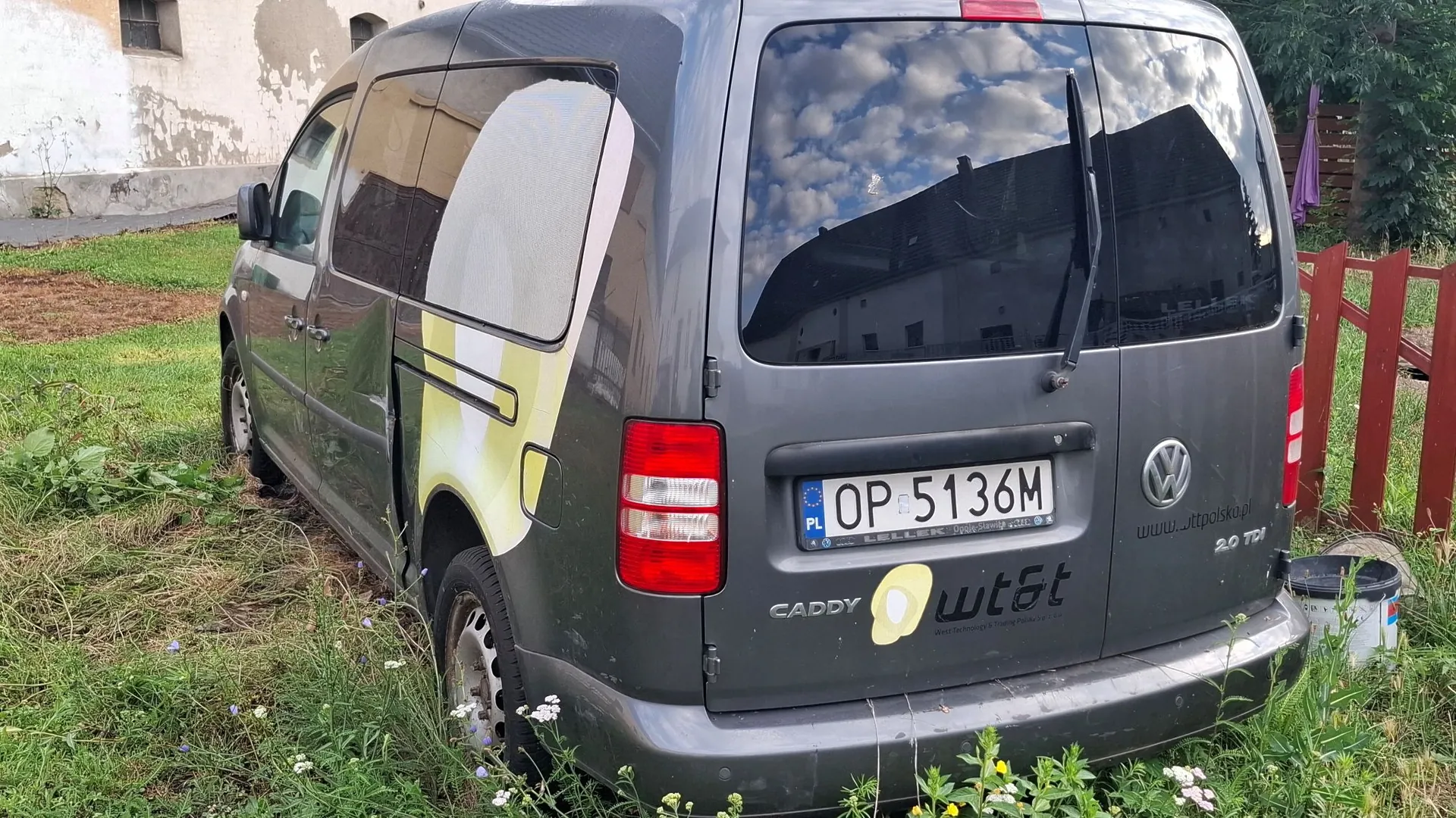 Srebrnoszary samochód dostawczy Volkswagen Caddy zaparkowany na trawie. Pojazd ma na boku i z tyłu żółto-białe grafiki oraz napis 'wt&t'. Z tyłu widoczna jest tablica rejestracyjna o numerze OP 5136M, logo Volkswagena oraz oznaczenie modelu 'CADDY' i silnika '2.0 TDI'. W tle widać fragment białego budynku oraz czerwony drewniany płot.