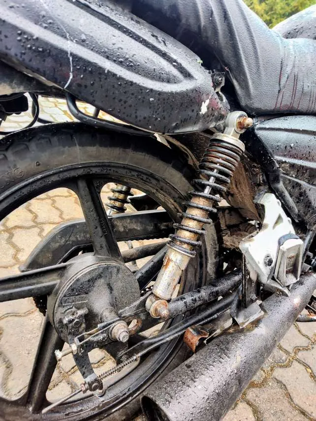 Zbliżenie na tylną część motocykla, pokazujące koło z oponą, zardzewiały amortyzator, elementy mechanizmu hamulcowego i łańcucha. Części motocykla są mokre, z widocznymi kroplami wody. Tło stanowi brukowana powierzchnia z geometrycznym wzorem. Widoczne są ślady zużycia i brudu na metalowych elementach.