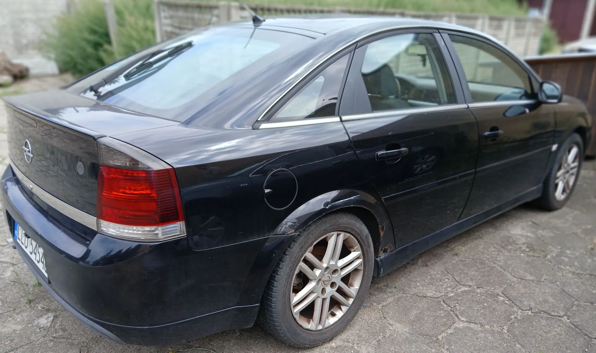 Czarny samochód osobowy typu sedan zaparkowany na kostce brukowej. Zdjęcie przedstawia tył i prawy bok pojazdu. Widoczna jest tylna lampa, klapa bagażnika z logo marki Opel oraz fragment tablicy rejestracyjnej. Nadwozie posiada widoczne ślady korozji nad tylnym prawym kołem.