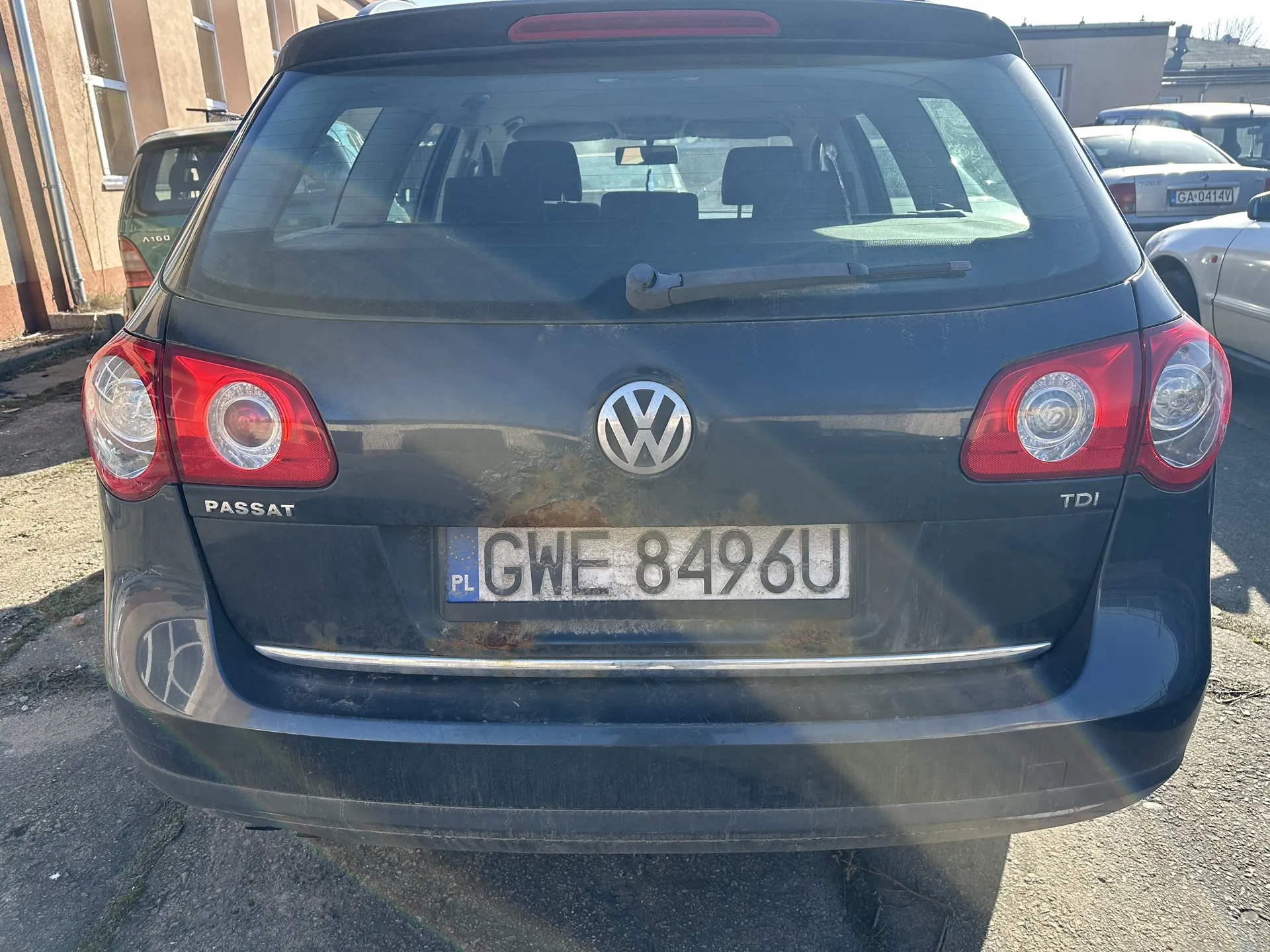 Tylne widok ciemnoszarego samochodu Volkswagen Passat TDI z tablicą rejestracyjną PL GWE 8496U. Widoczne są czerwone światła tylne, logo VW oraz napisy PASSAT i TDI. Na tylnej części pojazdu widoczne ślady brudu i rdzy. Samochód zaparkowany na otwartym parkingu z innymi pojazdami i budynkami w tle.