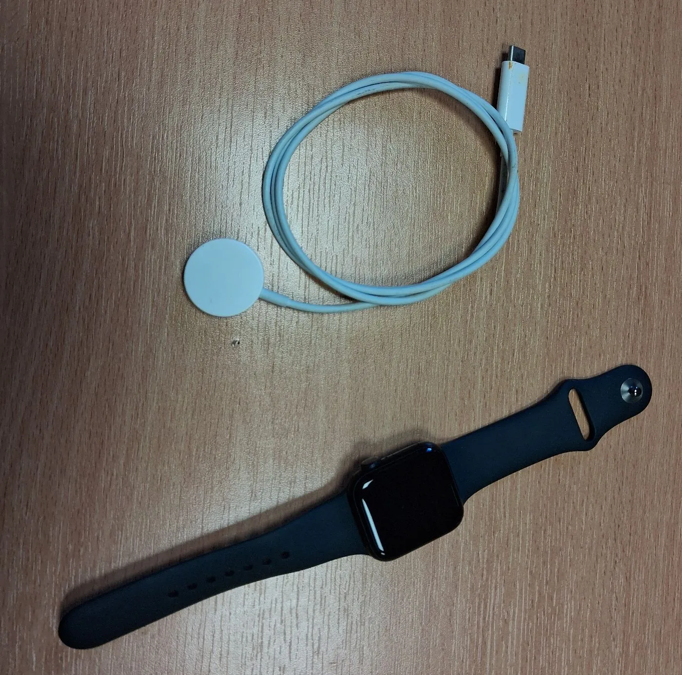 Na drewnianym blacie leży czarny smartwatch z gumowym paskiem oraz biały kabel do ładowania z magnetyczną końcówką w formie małego białego krążka i wtyczką USB.