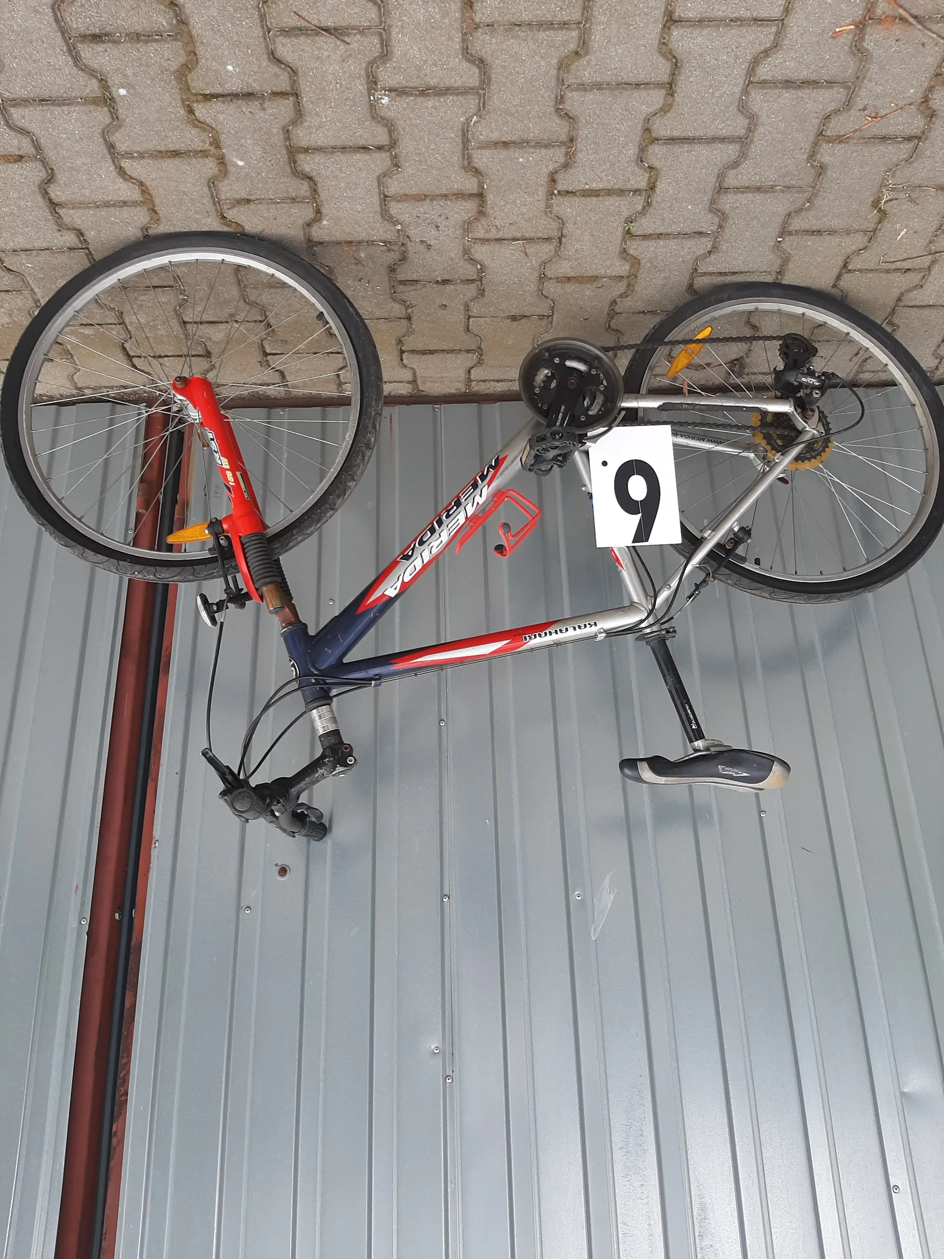 Rower o niebiesko-czerwonej ramie z białymi akcentami oparty o szarą metalową bramę garażową z pionowymi liniami. Na ramie widoczne napisy 'KALAHARI' i 'MERIDA'. Przy ramie umocowany biały znak z czarną cyfrą '9'. Tło stanowi brukowana powierzchnia.