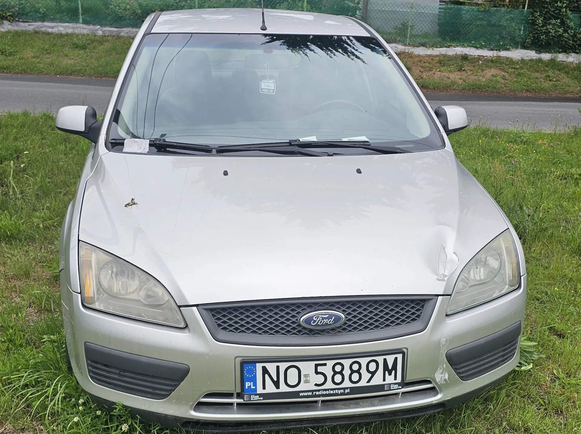 Srebrny samochód marki Ford widziany od przodu, zaparkowany na trawie obok drogi. Pojazd posiada wgniecenie na masce po prawej stronie oraz tablicę rejestracyjną o numerze NO 5889M z oznaczeniem PL i adresem strony internetowej www.radiooleszyn.pl.