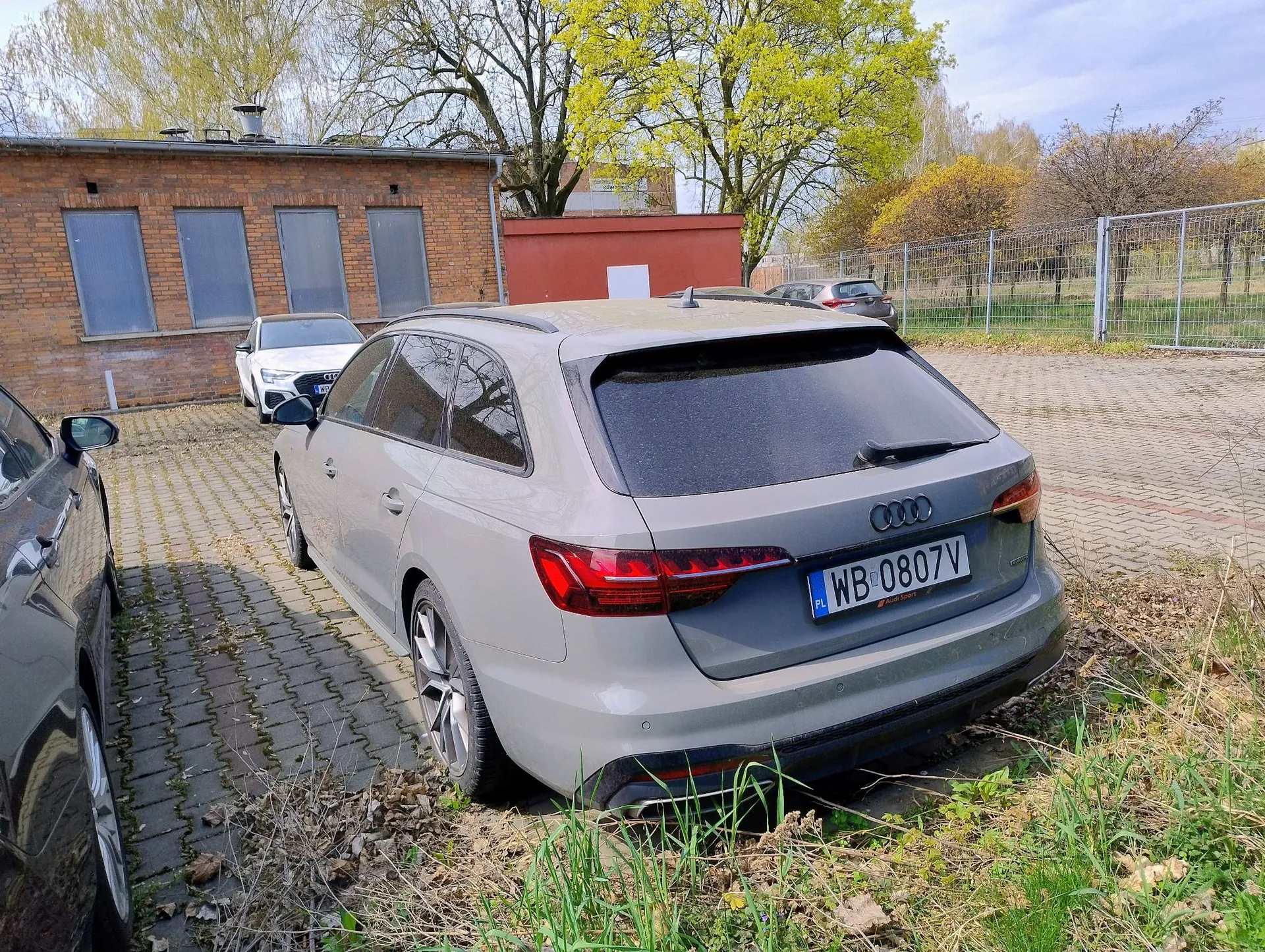 Szary samochód marki Audi w nadwoziu kombi zaparkowany na kostce brukowej. Na tablicy rejestracyjnej widnieje napis WB 0807V. W tle znajduje się ceglany budynek, czerwona ściana oraz drzewa z wiosennymi liśćmi. Po lewej stronie widoczny jest fragment innego, ciemnego samochodu.