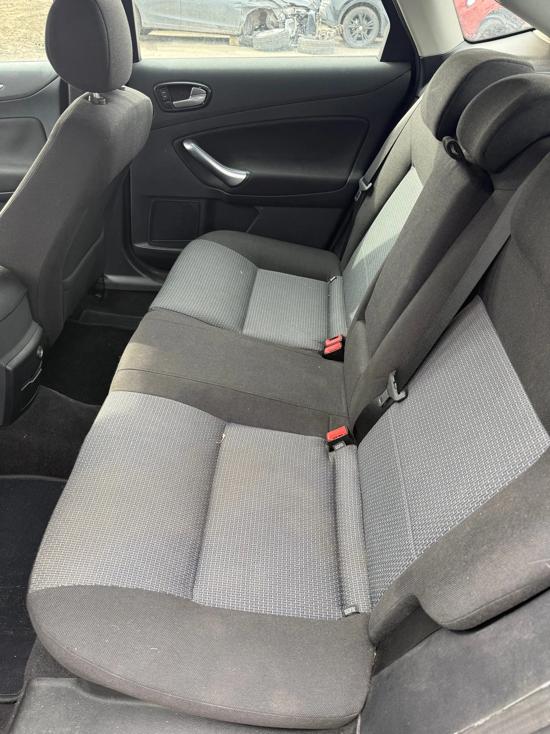 Wnętrze samochodu z widokiem na tylne siedzenia. Siedzenia tapicerowane szarym materiałem z geometrycznym wzorem. Widoczne pasy bezpieczeństwa z czerwonymi zapięciami. Drzwi z czarną obudową, uchwytem i okienkiem. Przez przednią szybę widoczny zewnętrzny widok z innym samochodem i oponami na tle.