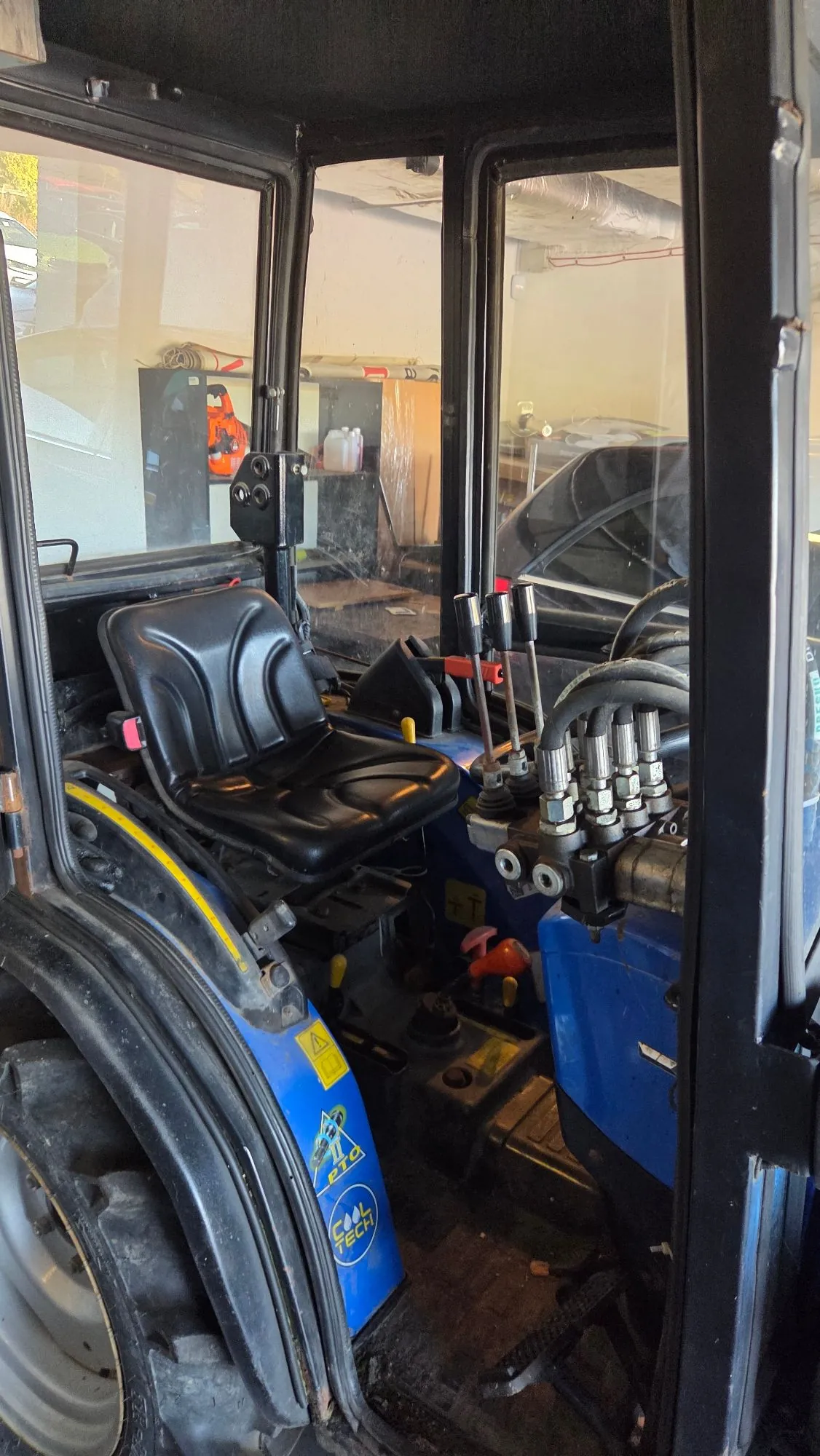 Wnętrze kabiny niebieskiego ciągnika rolniczego. Widoczny jest czarny, plastikowy fotel kierowcy, zestaw dźwigni sterujących oraz zawory hydrauliczne. Na niebieskim elemencie konstrukcyjnym znajdują się żółte tabliczki ostrzegawcze oraz logo 'COOL TECH'. W tle, za szybą kabiny, widoczny jest fragment warsztatu z narzędziami i sprzętem.