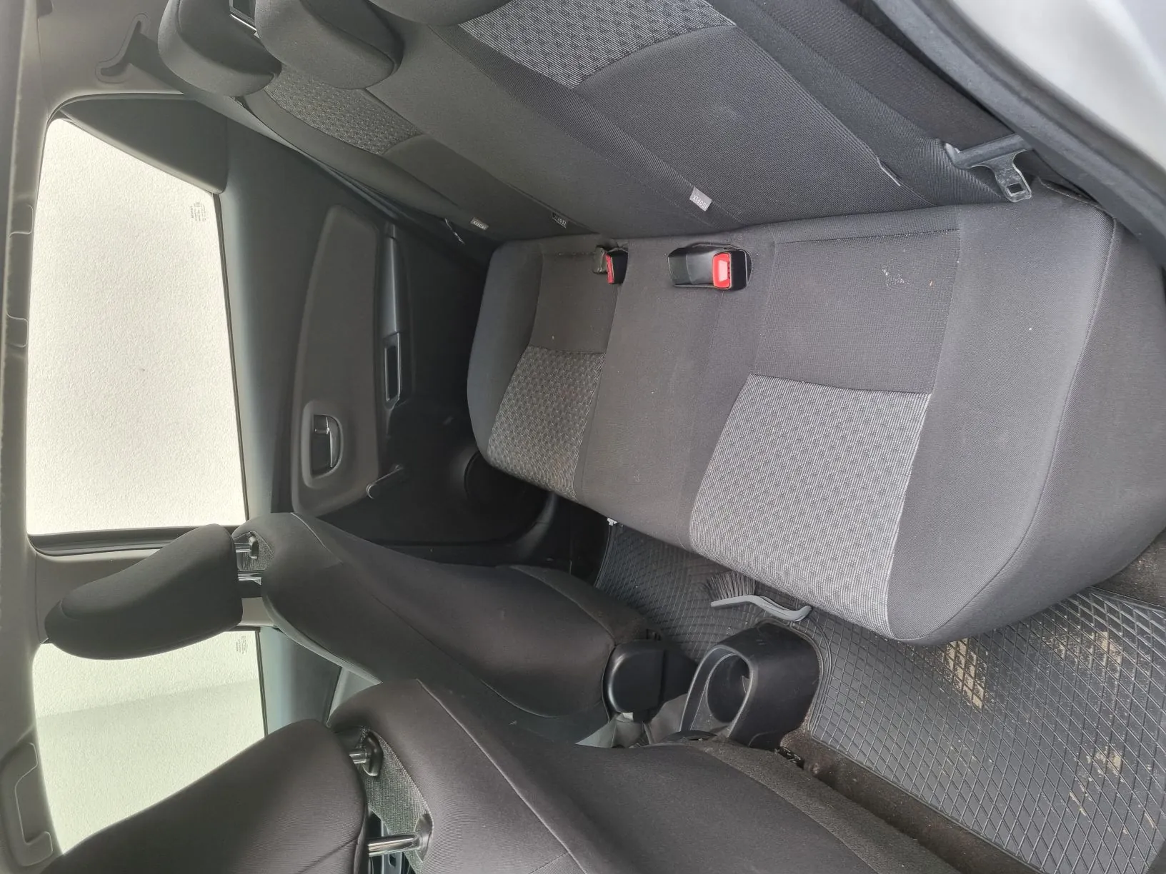 Wnętrze samochodu z widokiem na tylną kanapę. Szare siedzenia z tkaniną w geometryczny wzór, czerwone klamry pasów bezpieczeństwa. Czarna przednia kanapa częściowo widoczna. Drzwi z oknem i uchwytem po lewej stronie. Gumowe dywaniki podłogowe z widocznymi zabrudzeniami.