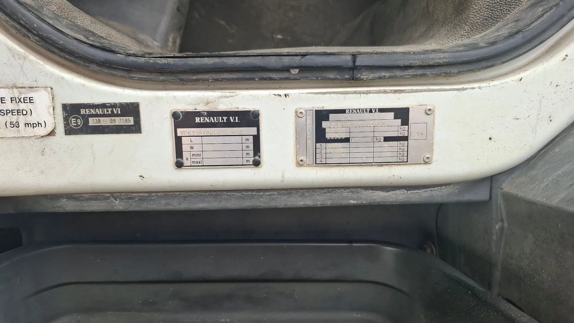 Zbliżenie na białą powierzchnię pojazdu z trzema metalowymi tabliczkami znamionowymi. Pierwsza tabliczka po lewej zawiera napis 'RENAULT VI' oraz oznaczenie 'E9 138-H 09 7185'. Środkowa tabliczka z napisem 'RENAULT V.I.' zawiera numer seryjny 'VF633AVAV0000104X58' oraz tabelę z parametrami L, W, mini, maxi w metrach. Trzecia tabliczka po prawej, również z napisem 'RENAULT V.I.', zawiera dane techniczne dotyczące masy w kilogramach (kg).