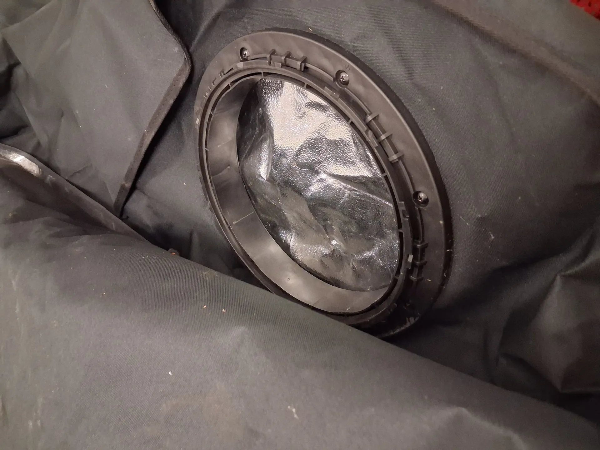 Zbliżenie na okrągły otwór w czarnym, materiałowym pokrowcu. Otwór jest oprawiony w czarną, plastikową ramkę z widocznymi śrubami mocującymi. Wewnątrz otworu znajduje się srebrzysta, refleksyjna folia aluminiowa.