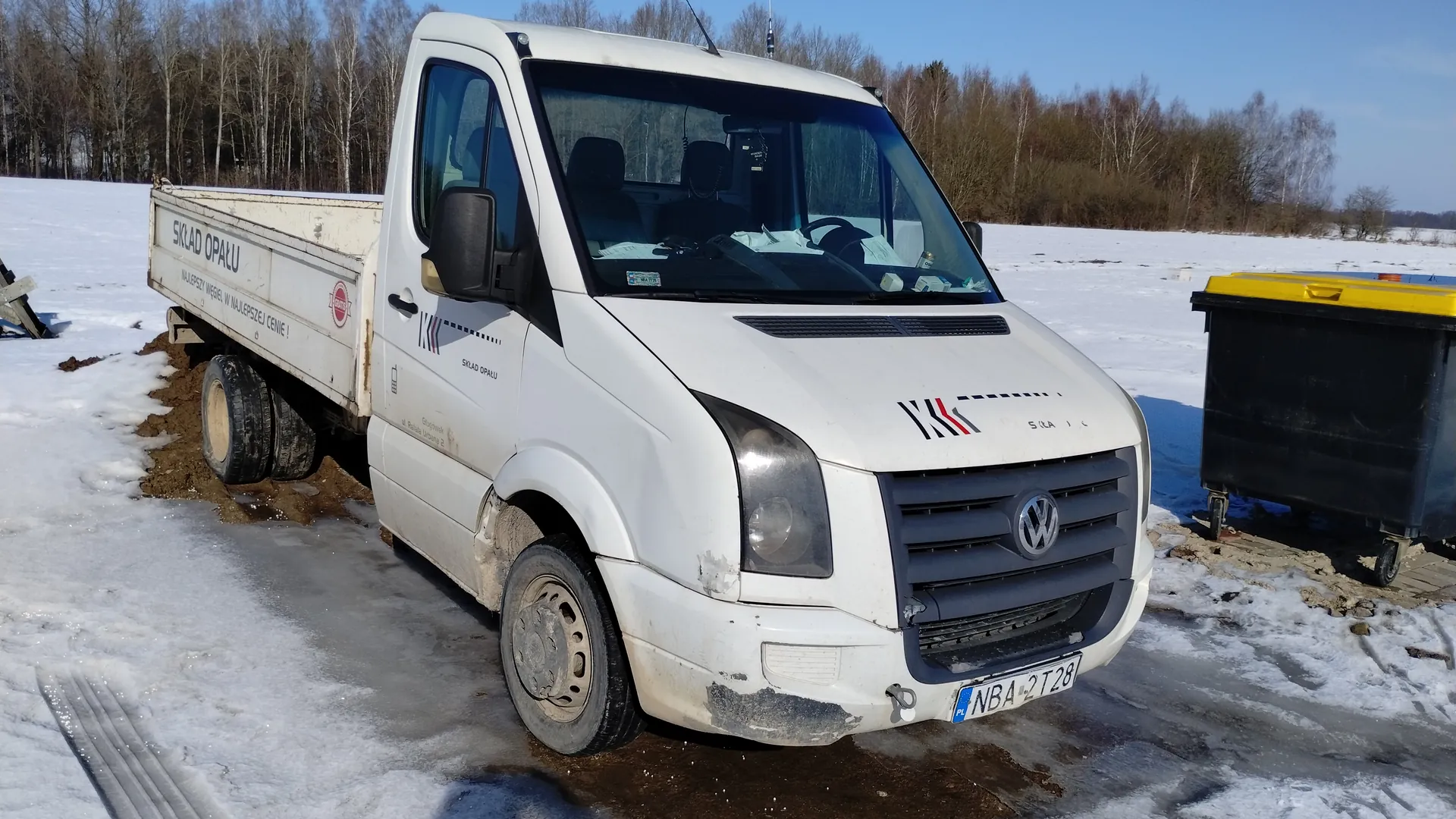 Biały samochód dostawczy marki Volkswagen z napisem 'SKŁAD OPAŁU' oraz 'NAJLEPSZY WĘGIEL W NAJLEPSZEJ CENIE!' na bocznej ścianie przyczepy. Na masce widoczne logo 'KK' z czerwonym i czarnym akcentem oraz tablica rejestracyjna 'NBA 2T28'. Pojazd stoi na śnieżnej powierzchni z odsłoniętymi plamami ziemi, obok czarnego kontenera z żółtym wiekiem. W tle las bez liści i jasne niebo. Samochód ma widoczne ślady zużycia i brudu na karoserii.
