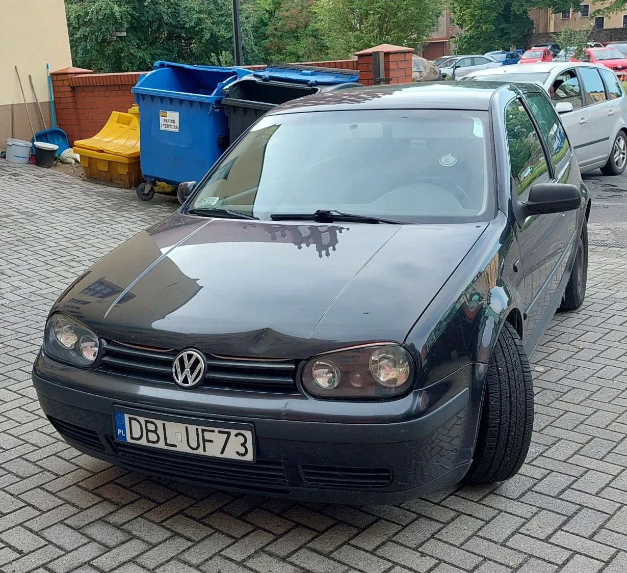 Czarny samochód marki Volkswagen Golf zaparkowany na kostce brukowej. Pojazd jest ustawiony przodem do widza, widoczna jest tablica rejestracyjna o numerze DBL UF73. W tle znajduje się niebieski kontener na odpady z napisem 'PAPIER I TEKTURA', żółty pojemnik oraz fragmenty budynków i zieleni.