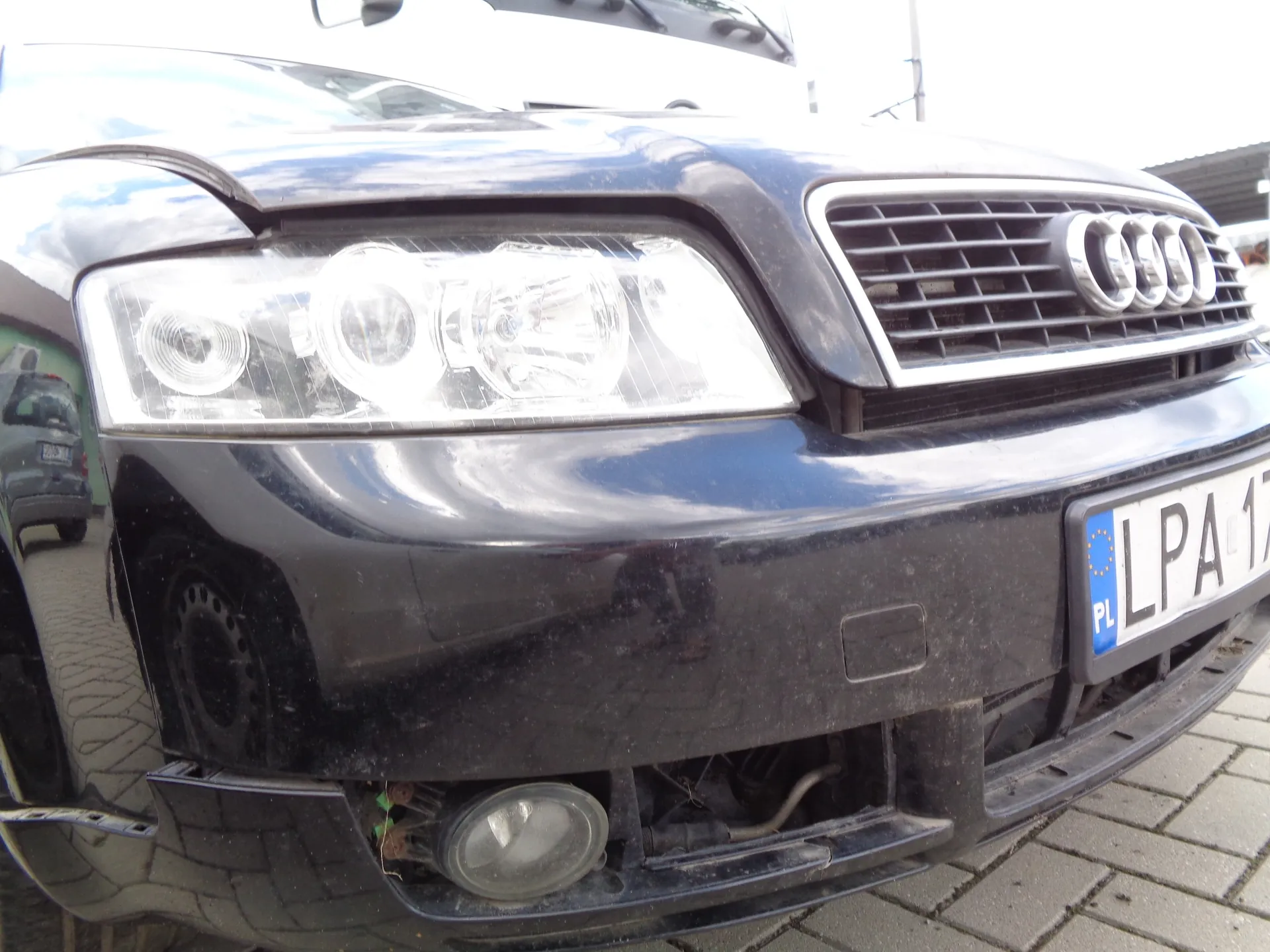 Zbliżenie na przód czarnego samochodu marki Audi. Widoczny jest prawy reflektor, grill z logo czterech pierścieni oraz fragment tablicy rejestracyjnej z oznaczeniem PL i literami LPA. Samochód znajduje się na kostce brukowej.
