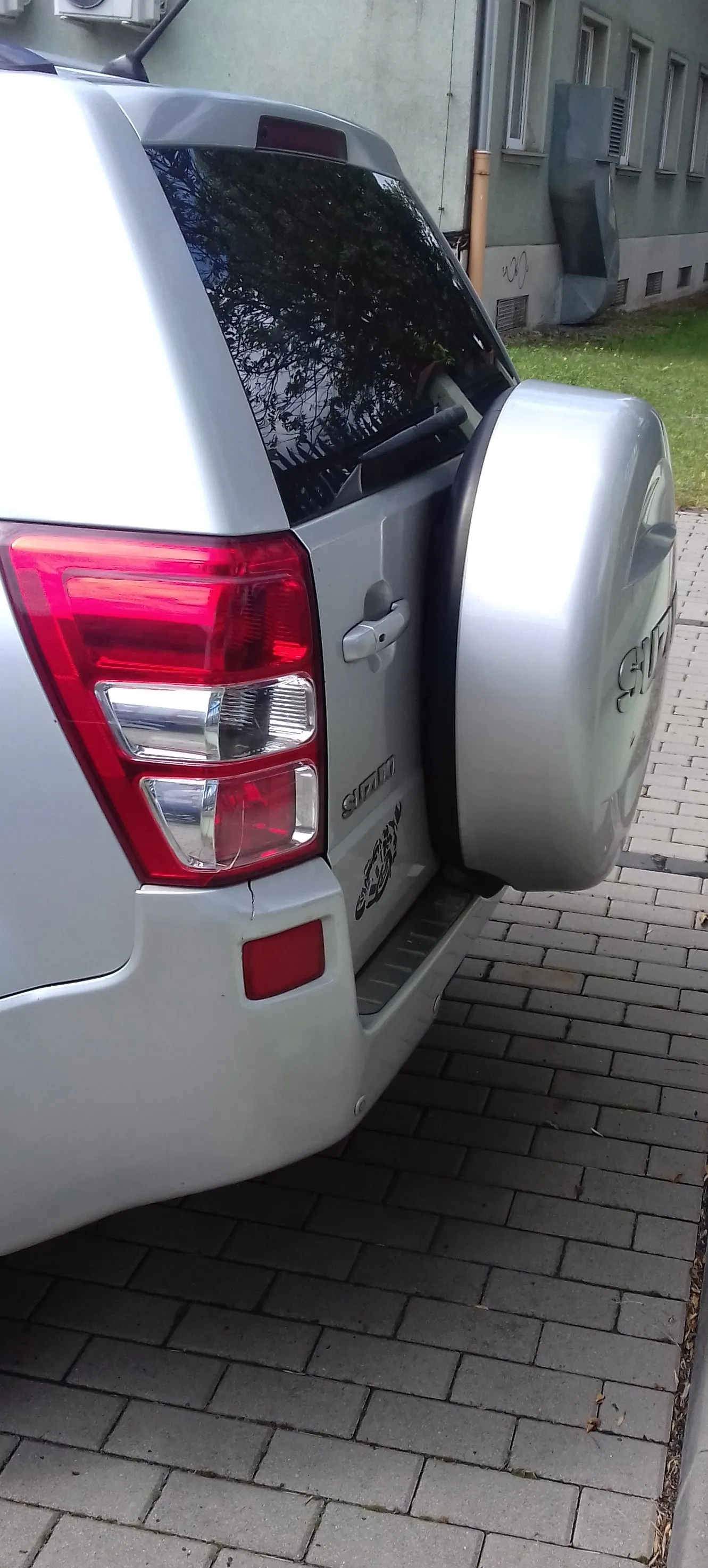 Tylna część srebrnego samochodu typu SUV zaparkowanego na kostce brukowej. Widoczny jest lewy tylny reflektor, tylna szyba oraz zapasowe koło z twardą, srebrną osłoną zamontowaną na klapie bagażnika. Na klapie znajduje się napis 'GRAND'. W tle widoczny jest fragment jasnego budynku i trawnik.