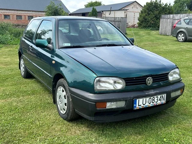 Ciemnozielony samochód osobowy marki Volkswagen Golf trzeciej generacji zaparkowany na trawniku. Pojazd ma czarne zderzaki i polską tablicę rejestracyjną o numerze LU 0834N. W tle widoczne są budynki gospodarcze oraz fragment innego, srebrnego samochodu.