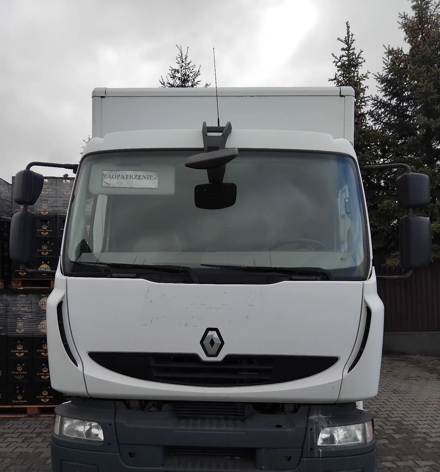 Widok z przodu białego samochodu dostawczego marki Renault. Na przedniej szybie, po lewej stronie, znajduje się biała kartka z napisem „ZAOPATRZENIE”. Pojazd ma duże lusterka boczne i ciemny grill z logo Renault na środku. W tle widoczne są ciemne skrzynie transportowe oraz drzewa iglaste pod zachmurzonym niebem.