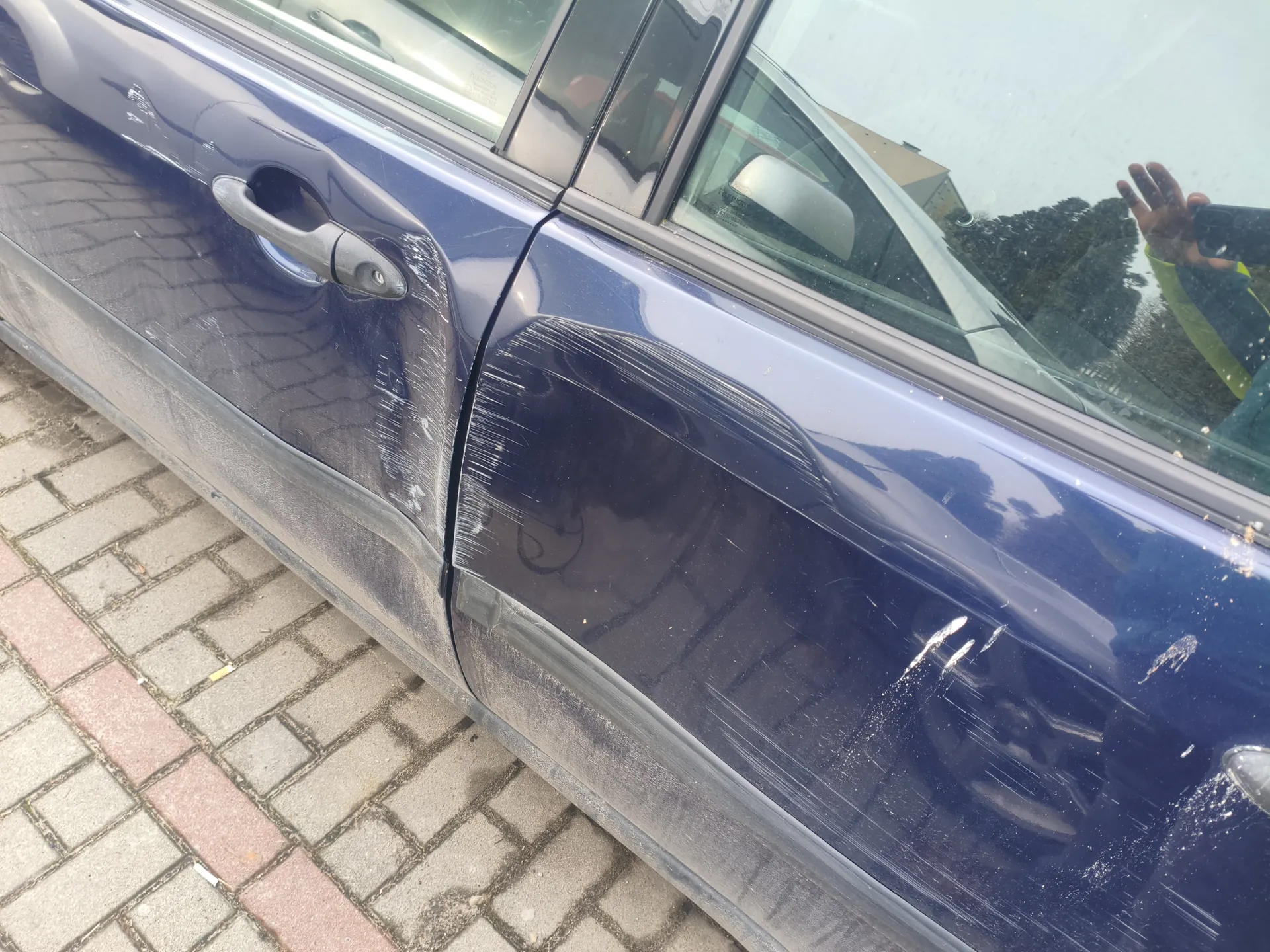 Boczny widok niebieskiego samochodu z widocznymi zadrapaniami na przedniej drzwi. Samochód stoi na brukowanej nawierzchni. W tle przez przednią szybę widoczna jest ręka osoby trzymającej telefon, a także odbicie drzew i budynku.