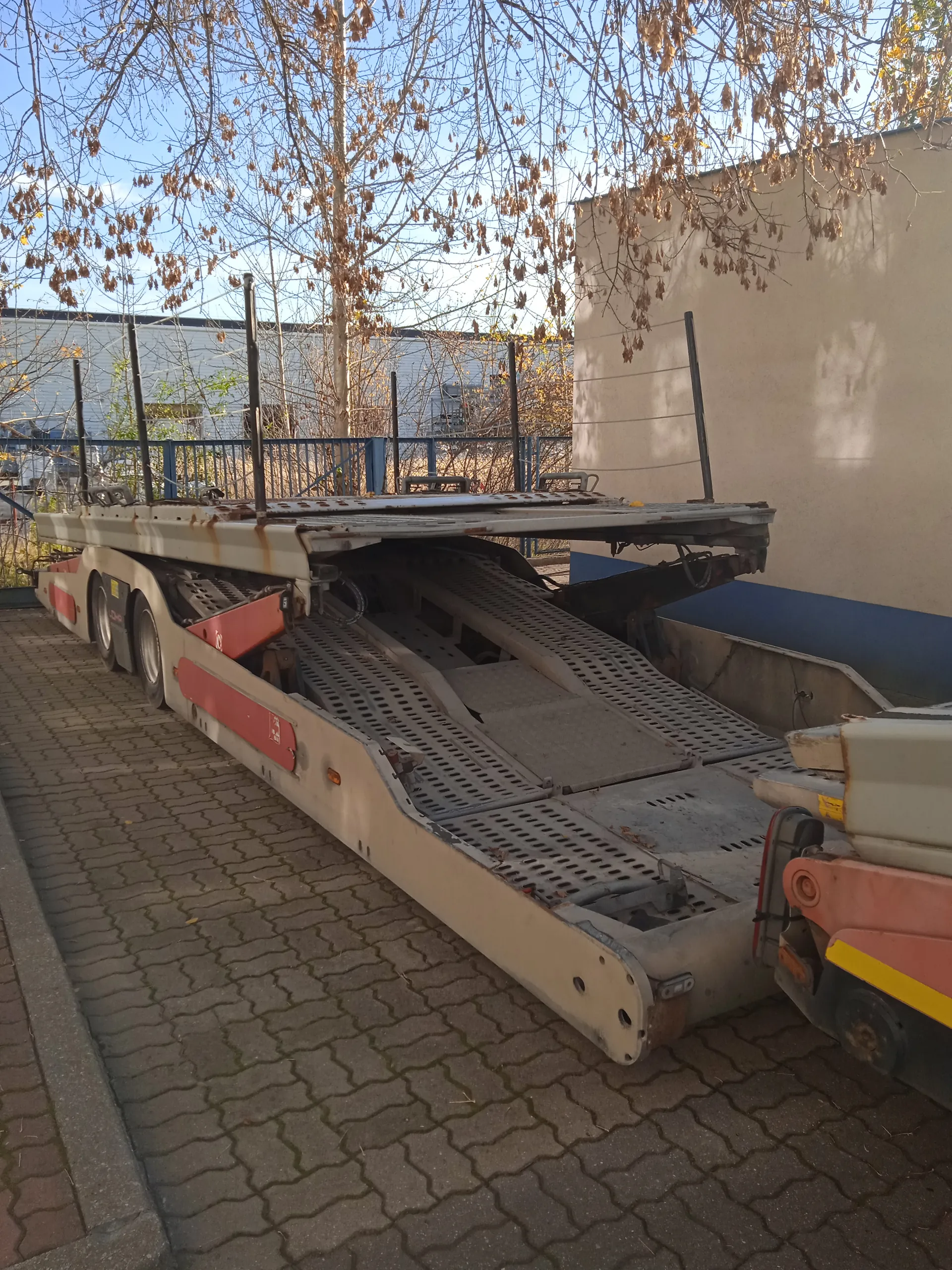 Przyczepa transportowa do przewozu samochodów z rozłożonym rampą, zaparkowana na brukowanej powierzchni. Przyczepa ma beżowo-czerwony kolor z widocznymi śladami korozji. Na boku przyczepy widoczny napis '24'. W tle budynek o beżowo-niebieskiej elewacji, drzewa bez liści i niebo. Na przyczepie umieszczone są pionowe słupki oraz metalowe elementy konstrukcyjne.