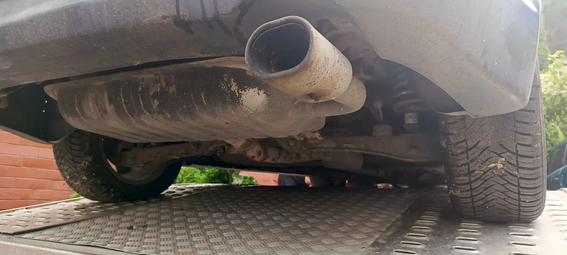 Zbliżenie na tylną część podwozia samochodu, ukazujące rurę wydechową z tłumikiem. Pojazd znajduje się na metalowej platformie transportowej o antypoślizgowej strukturze. Widoczne są również tylne opony oraz fragmenty zawieszenia.