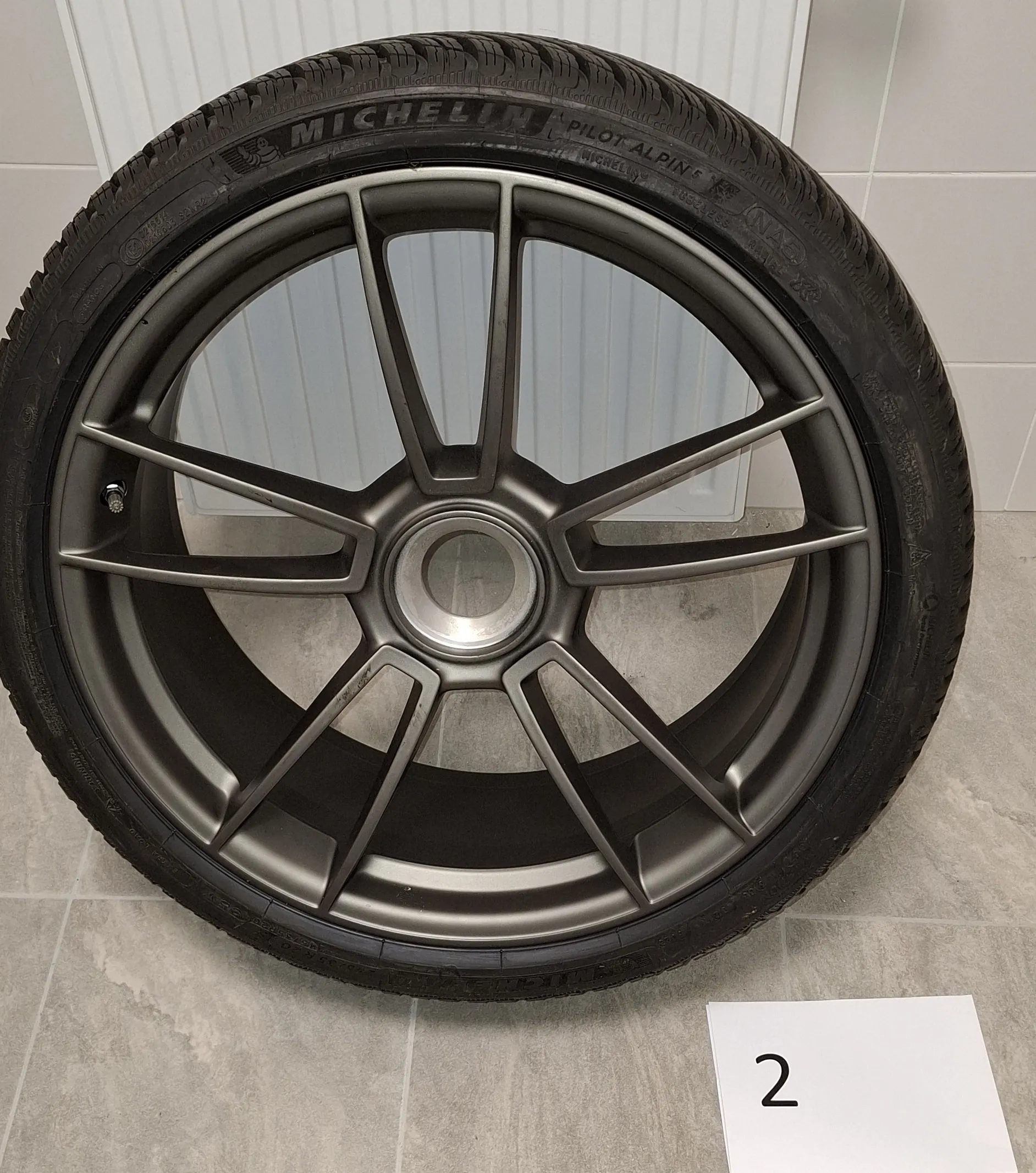 Zdjęcie koła samochodowego z oponą marki Michelin Pilot Alpine, zamontowanego na ciemnoszarej, matowej feldze aluminiowej o pięciu podwójnych ramionach. Koło znajduje się na tle białego grzejnika i jasnoszarej podłogi z płytek. W prawym dolnym rogu widoczna jest biała kartka z czarną cyfrą 2.