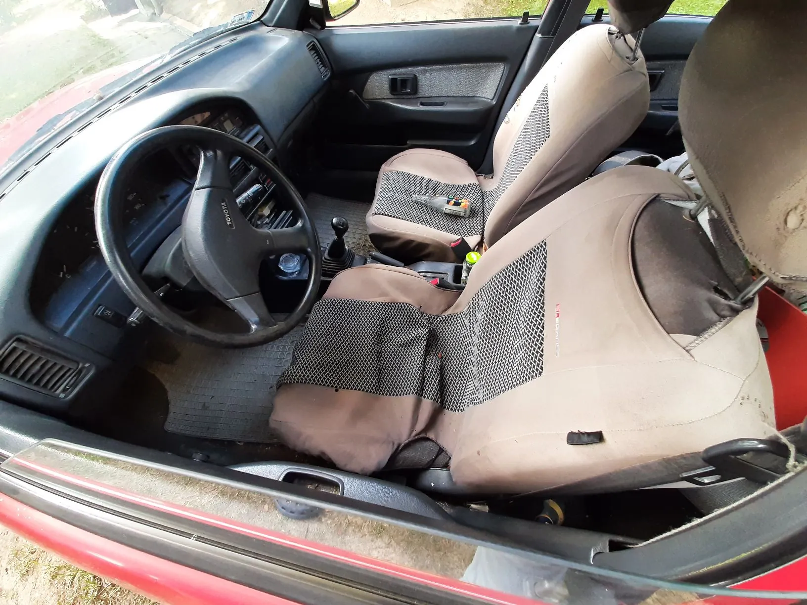 Wnętrze samochodu marki Toyota widziane z perspektywy otwartych drzwi kierowcy. Widoczna jest kierownica z logo Toyota, deska rozdzielcza, dwa przednie fotele z beżowymi pokrowcami z siatką oddychającą oraz fragment czerwonego nadwozia auta.