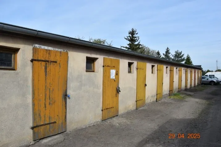 Długi, niski budynek gospodarczy o szarych ścianach z rzędem żółtych, drewnianych drzwi. Pomiędzy drzwiami znajdują się małe, kwadratowe okna. Przed budynkiem rozciąga się utwardzona droga z ziemi i żwiru. W tle widoczne są drzewa iglaste oraz zaparkowane samochody. W prawym dolnym rogu zdjęcia znajduje się czerwony napis z datą: 29.04.2025.