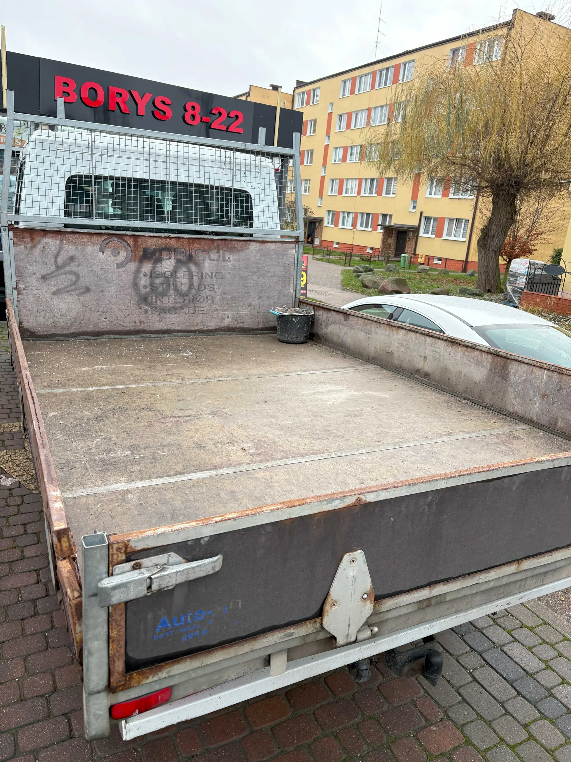 Zdjęcie przedstawia tylną część otwartego naczepa ciężarówki z metalowym ogrodzeniem. Na tle widoczny biały van z czerwonym napisem "BORYS 8-22" na czarnym tle. Na ścianie naczepa widoczne są napisy: "BORISOL", "ISOLERING", "STILLADS", "INTERIOR", "FACADE" oraz graffiti. W dolnej części naczepa widoczny niebieski napis "Auto" z dodatkowym, nieczytelnym tekstem. W tle żółty blok mieszkalny z czerwonymi akcentami, drzewo, srebrny samochód osobowy i czarny wiadro w naczepie. Podłoga wyłożona ceglanymi płytami. Niebo zachmurzone.