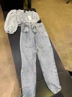 Jasnoniebieski dżinsowy kombinezon z podpaskami i białą etykietą na pasie, ułożony na ciemnej powierzchni. Tło stanowi żółtawa podłoga i fragment ciemnego mebla.