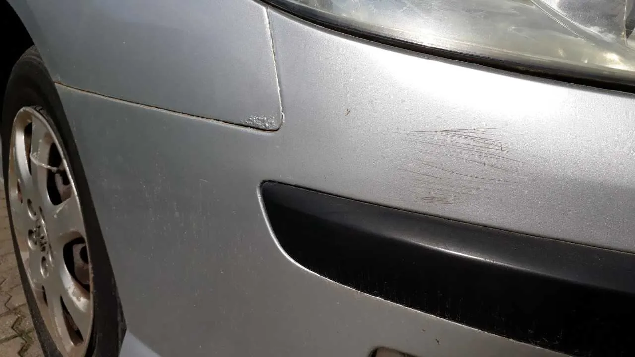 Zbliżenie na przód srebrnego samochodu. Widoczny jest reflektor, fragment koła z aluminiową felgą oraz zarysowana na błotniku rysa. Na karoserii znajdują się drobne zabrudzenia. W tle fragment chodnika z wzorzystą kostką.
