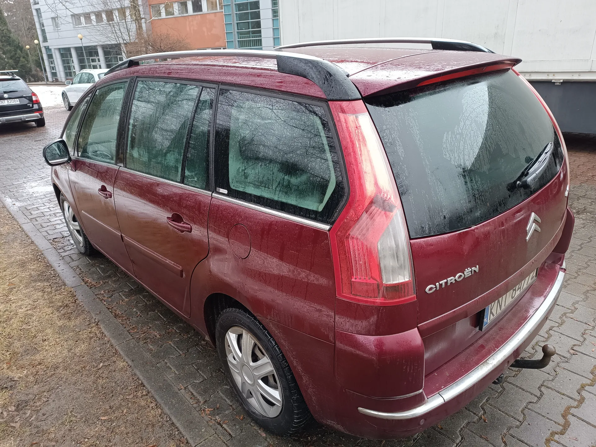 Czerwony samochód osobowy Citroën kombi z widocznym napisem "CITROËN" i tablicą rejestracyjną KNT 8477. Mokra, brudna karoseria, szyby z kroplami wody, dachówka. Zaparkowany na brukowanej powierzchni obok suchego trawnika, w tle budynki i inne pojazdy.