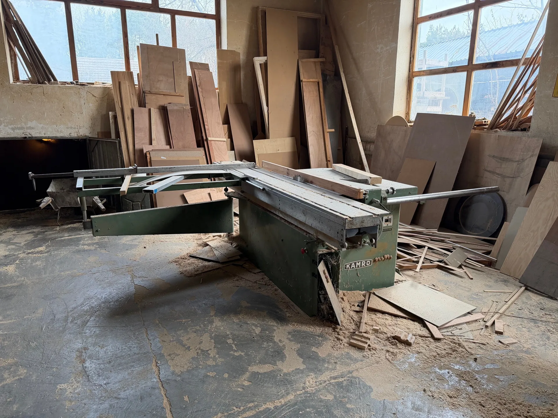 Wnętrze warsztatu z zielonym maszyną do obróbki drewna marki KAMRO umieszczoną w centrum. Wokół maszyny rozrzucone są deski, płyty drewniane i wióry. Na podłodze z betonu widoczne ślady pyłu i brudu. W tle duże okna z widokiem na zewnątrz, gdzie widać drzewa i budynek z blachą. Ściany pomieszczenia są zniszczone, a na maszynie widnieje napis KAMRO.