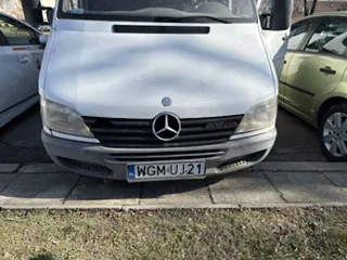 Przedni widok srebrnej furgonetki Mercedes-Benz z widocznym logo na masce i tablicą rejestracyjną WGM UJ 21. Po lewej stronie częściowo widoczny biały samochód, po prawej zielony samochód. Furgonetka zaparkowana na brukowanej powierzchni, na pierwszym planie trawa. Tło stanowi fragment budynku.