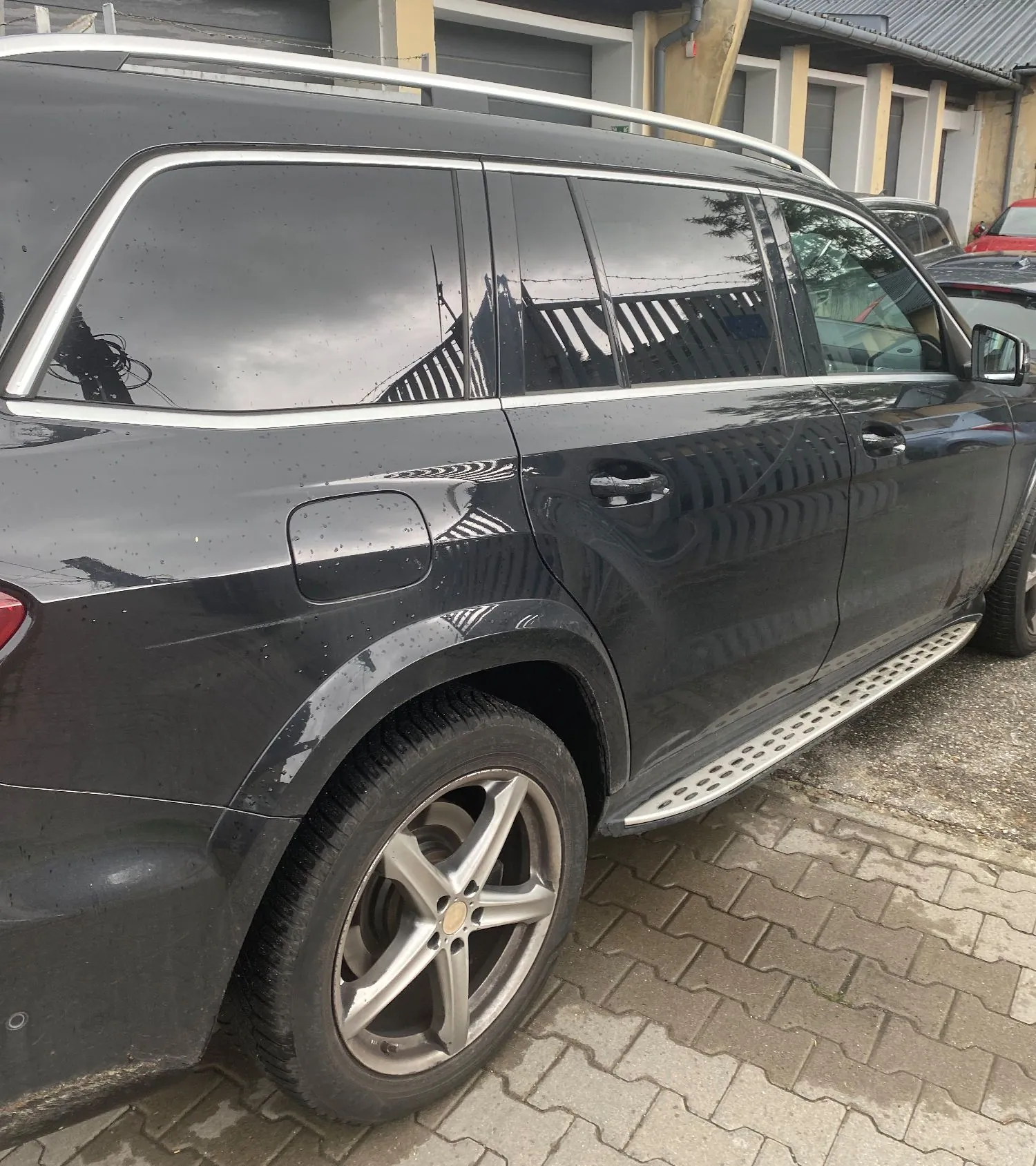 Zbliżenie na prawy bok czarnego samochodu typu SUV zaparkowanego na kostce brukowej. Widoczne są przyciemniane szyby, srebrne relingi dachowe oraz srebrny stopień boczny z perforacjami. Koło z pięcioramienną srebrną felgą jest skierowane lekko w lewo.